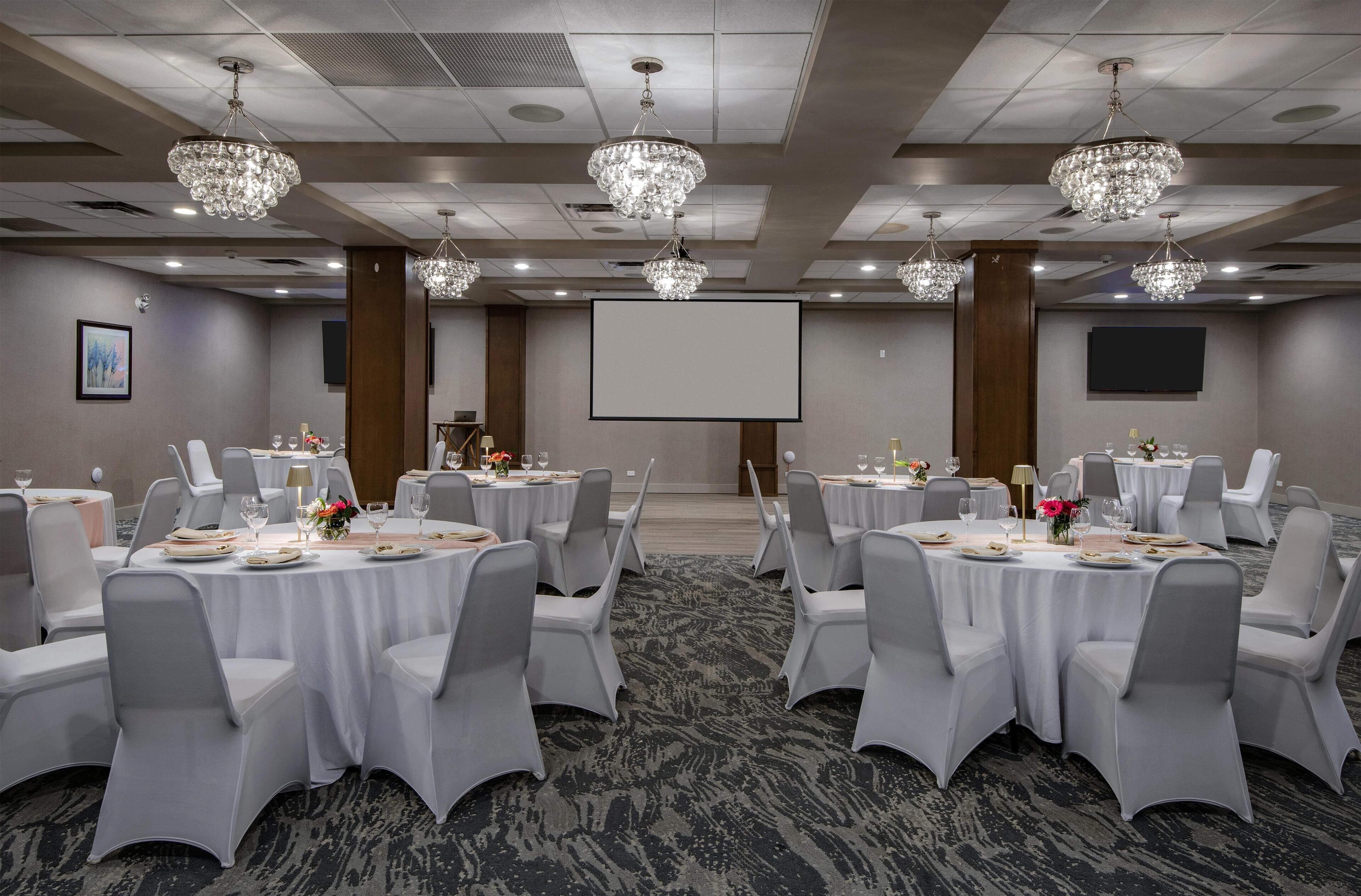 A conference room with round tables, white chairs, chandeliers, a projector screen, and a carpeted floor.