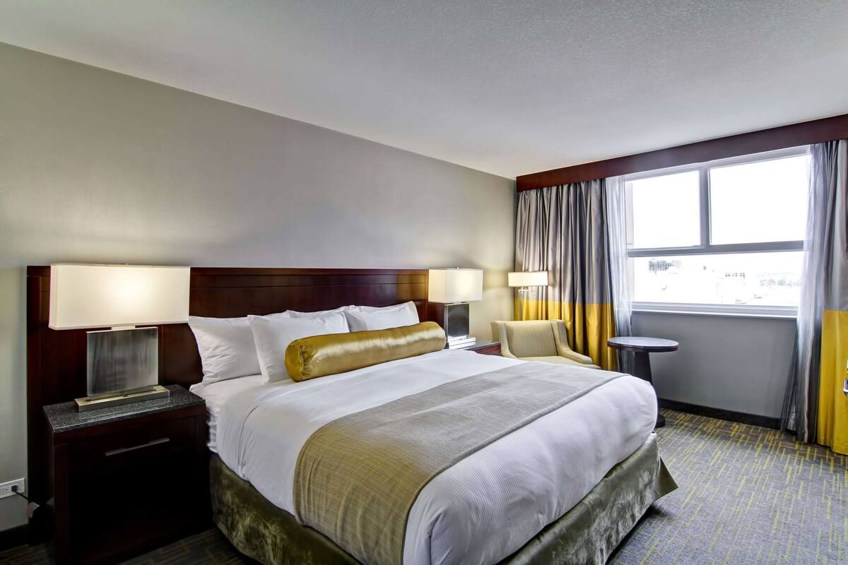 A hotel room with a large bed, bedside lamps, a small table, and a view of the outside through a window.
