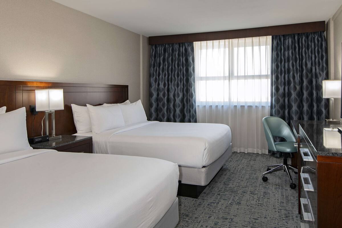 A hotel room with two beds, a desk with a chair, and a large window with curtains.