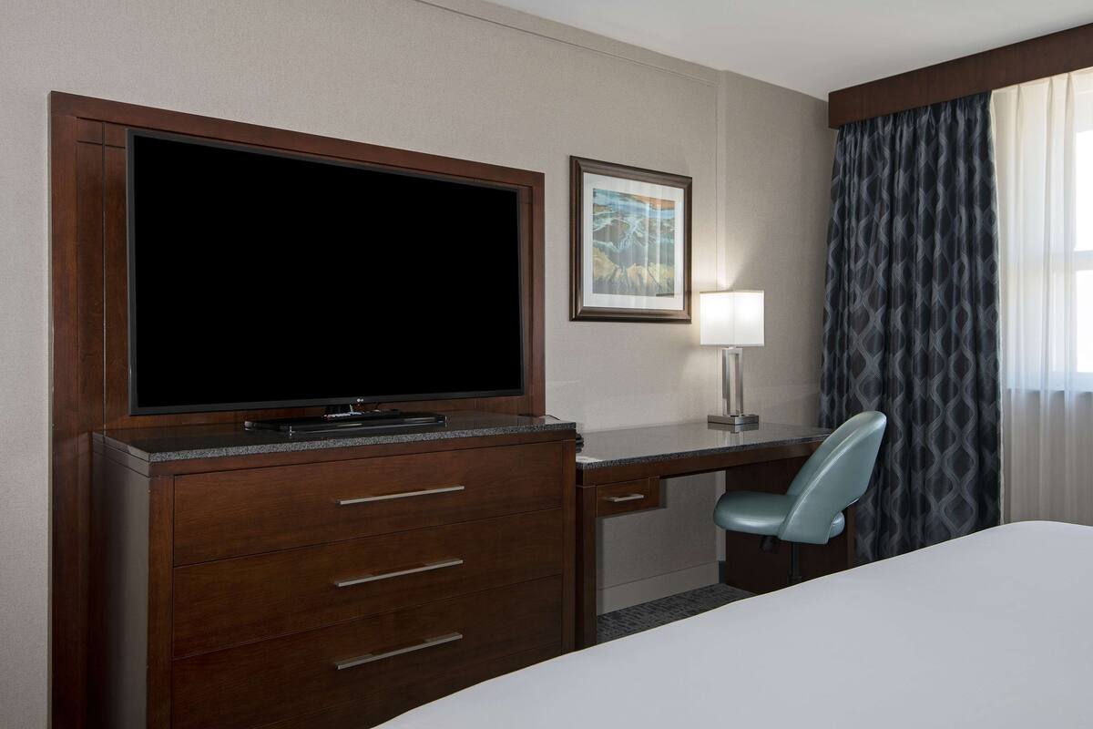 A hotel room with a large flat-screen TV, a desk with a chair, a bed, a framed picture on the wall, and a window with curtains.