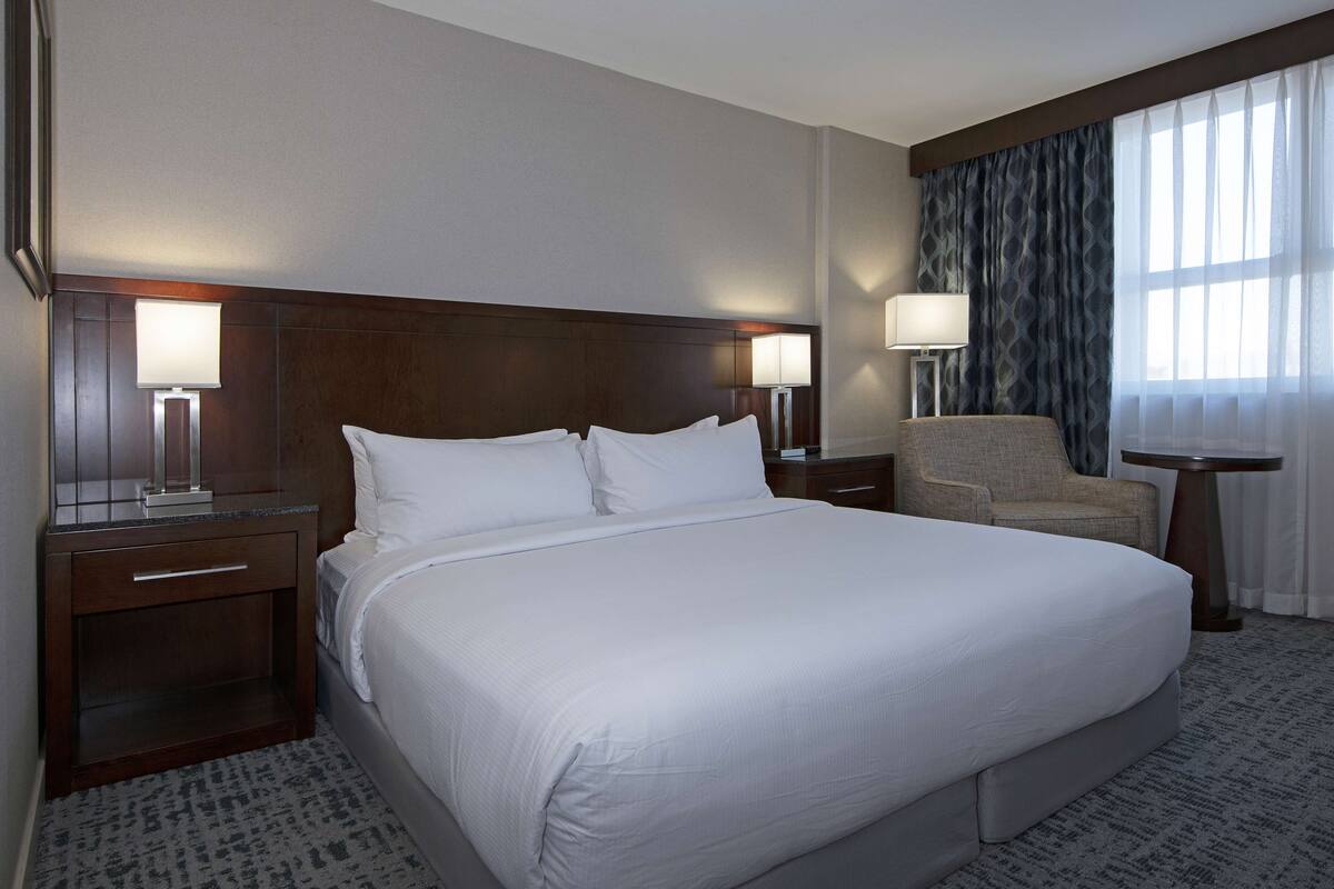 A hotel room with a large bed, two bedside tables with lamps, a chair, and a view of the outdoors through sheer curtains.