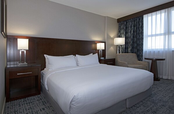 A hotel room with a large bed, two bedside tables with lamps, a chair, and a view of the outdoors through sheer curtains.