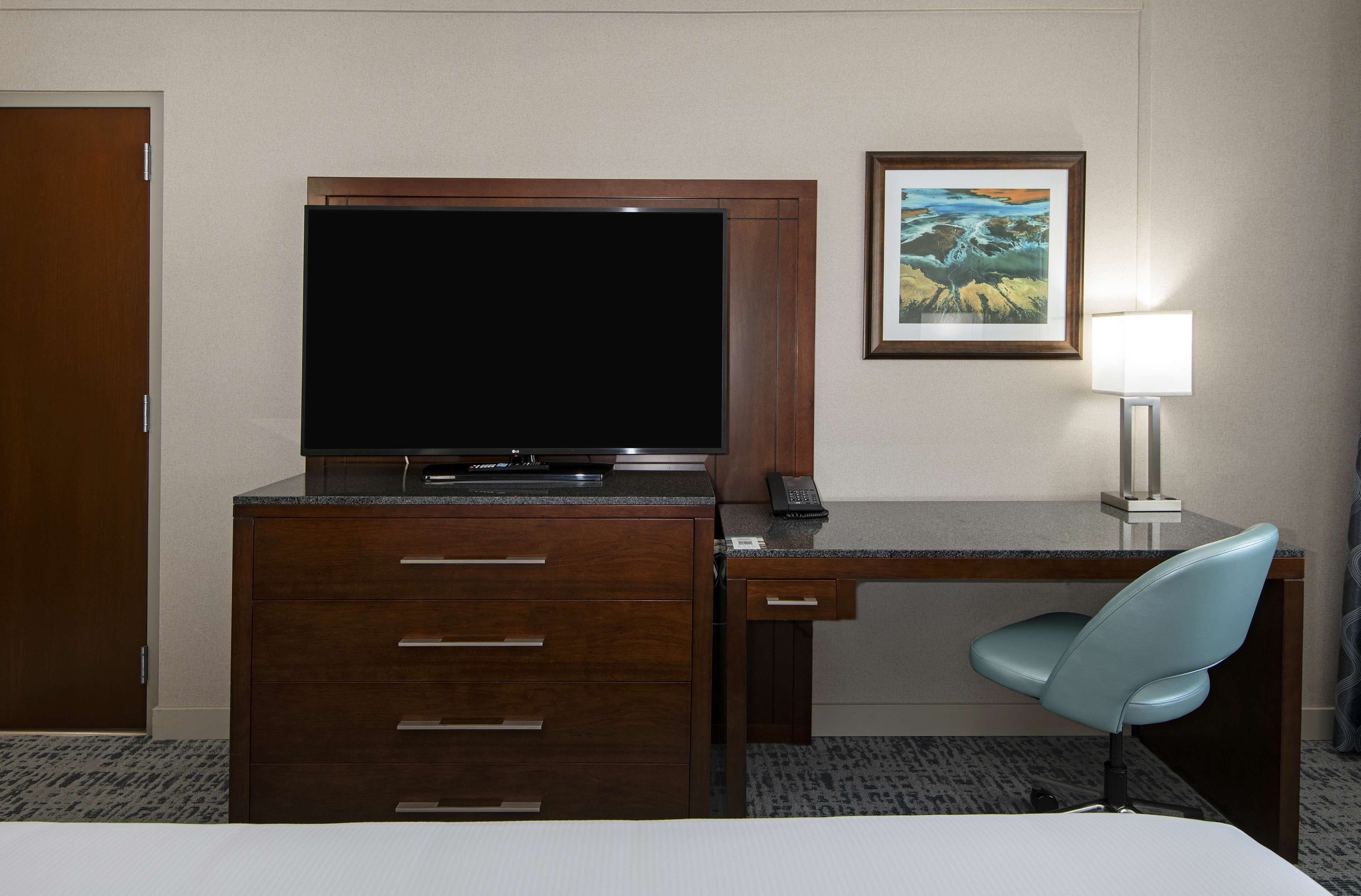 Habitació d'hotel amb escriptori de fusta, televisió de pantalla plana, làmpada de taula, quadre al mur i cadira blava.