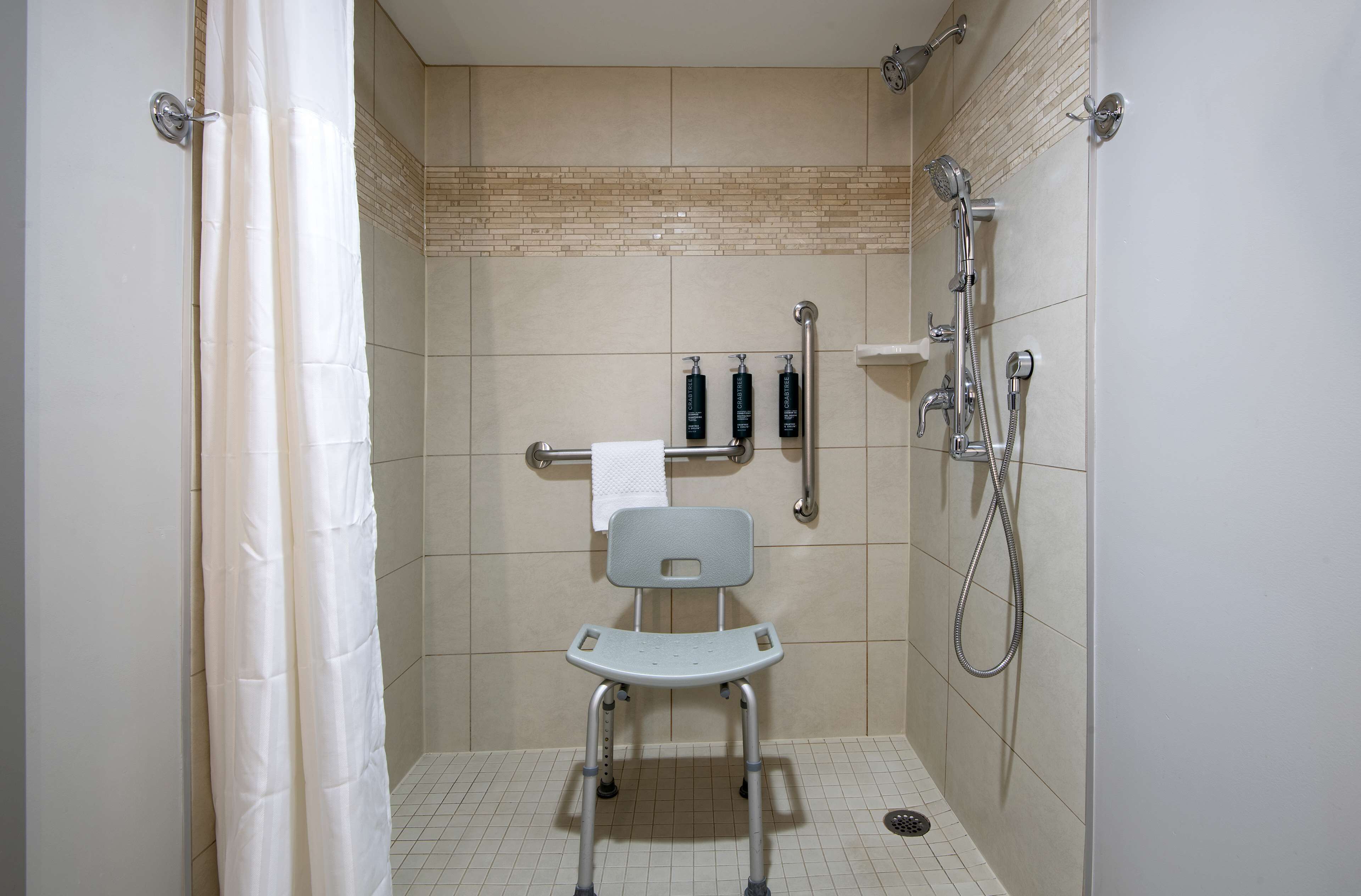 Un espace douche équipé d’un siège de douche accessible aux personnes en fauteuil roulant, de barres d’appui et d’un pommeau de douche à main.
