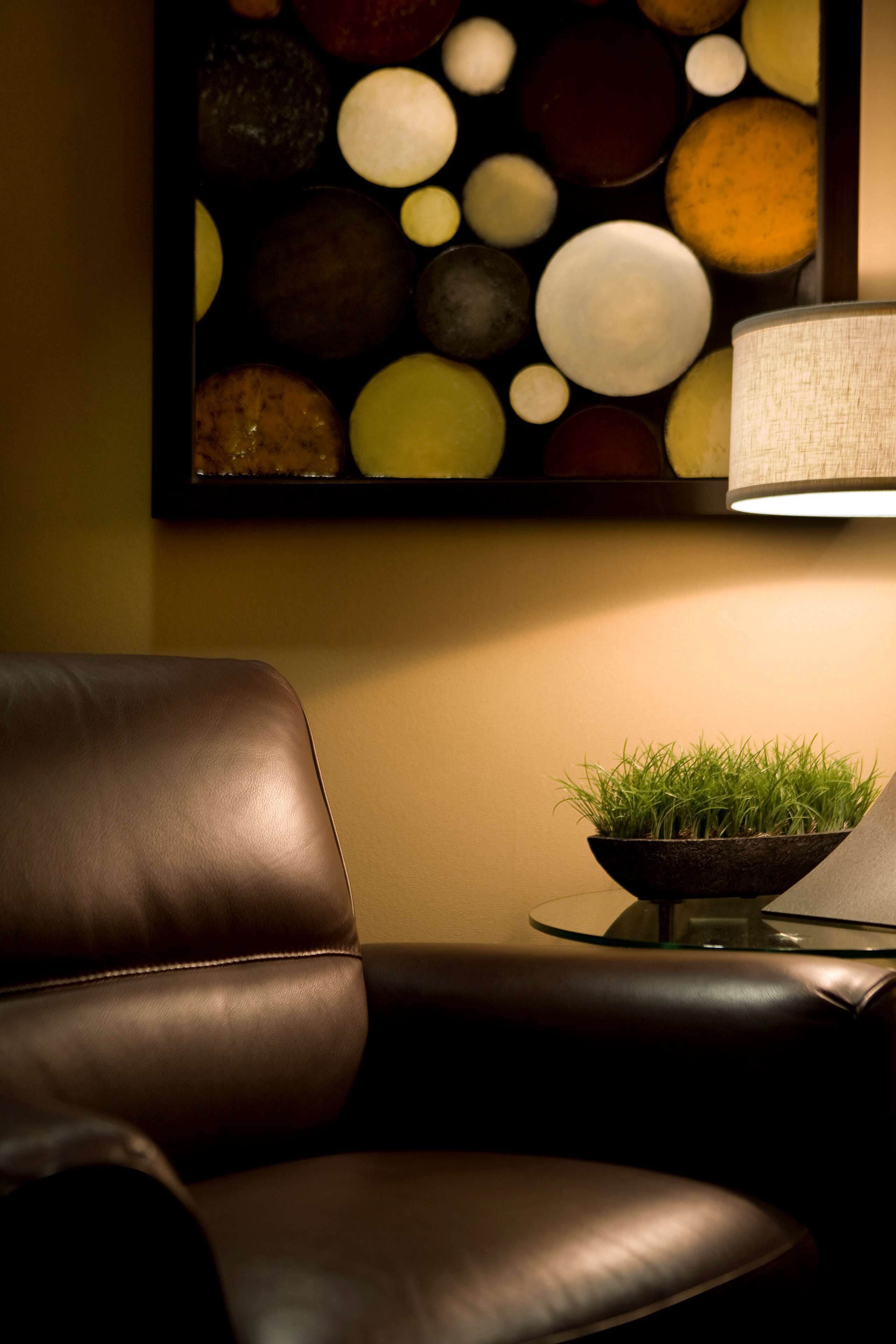 A leather chair, a round glass table with a bowl of green plants, and a wall-mounted abstract painting.