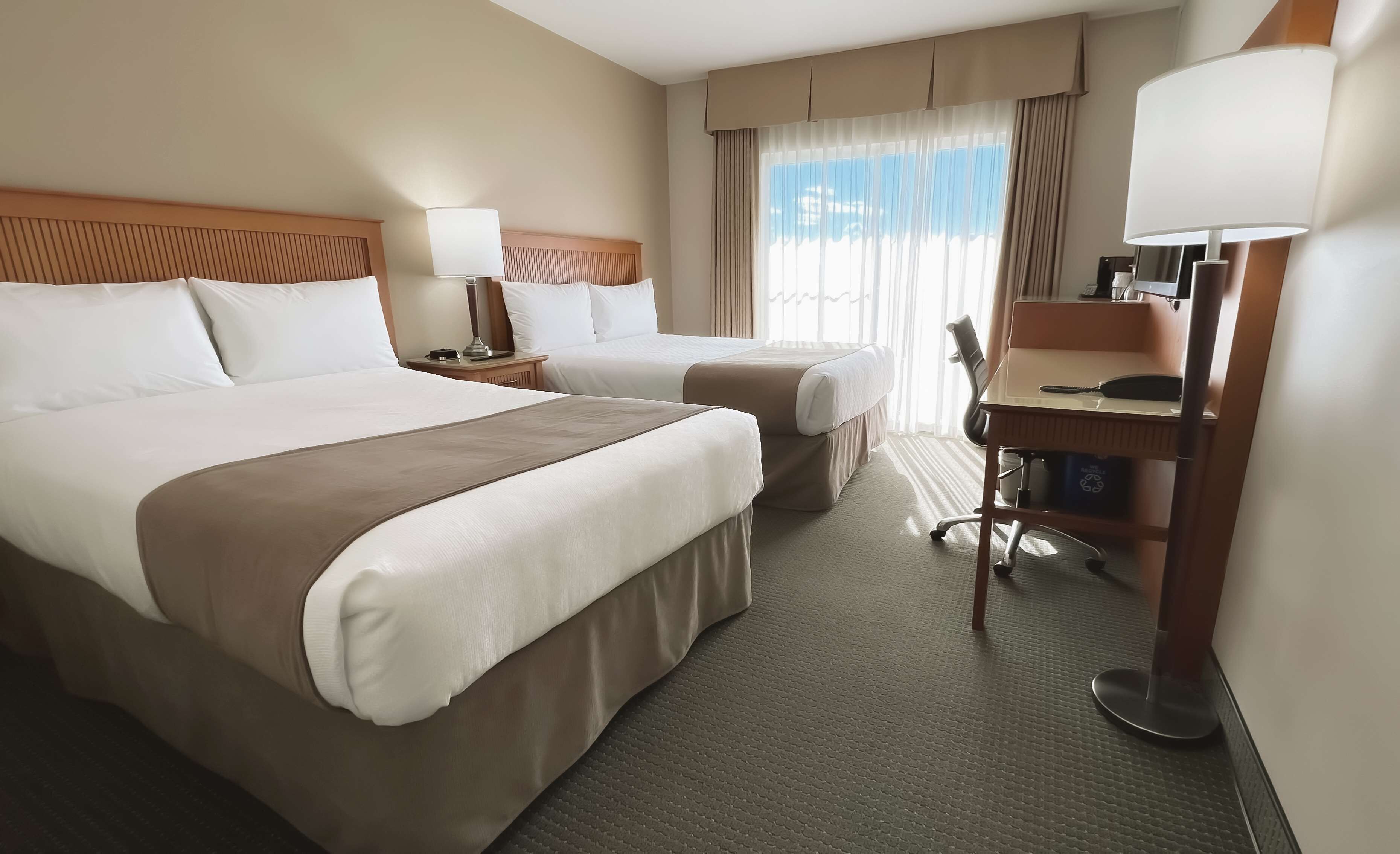Habitación de hotel con dos camas, un escritorio, una silla, una lámpara y un ventanal con cortinas.