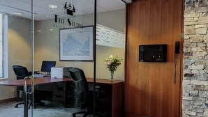 Un salón de conferencias con un escritorio de madera, sillas de oficina negras, un separador de vidrio, un cuadro enmarcado, un jarrón con flores, una pared de piedra y una puerta de madera.