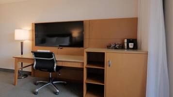 A hotel room with a wooden desk, a black office chair, a flat-screen TV mounted on the wall, a coffee maker, and a minibar.