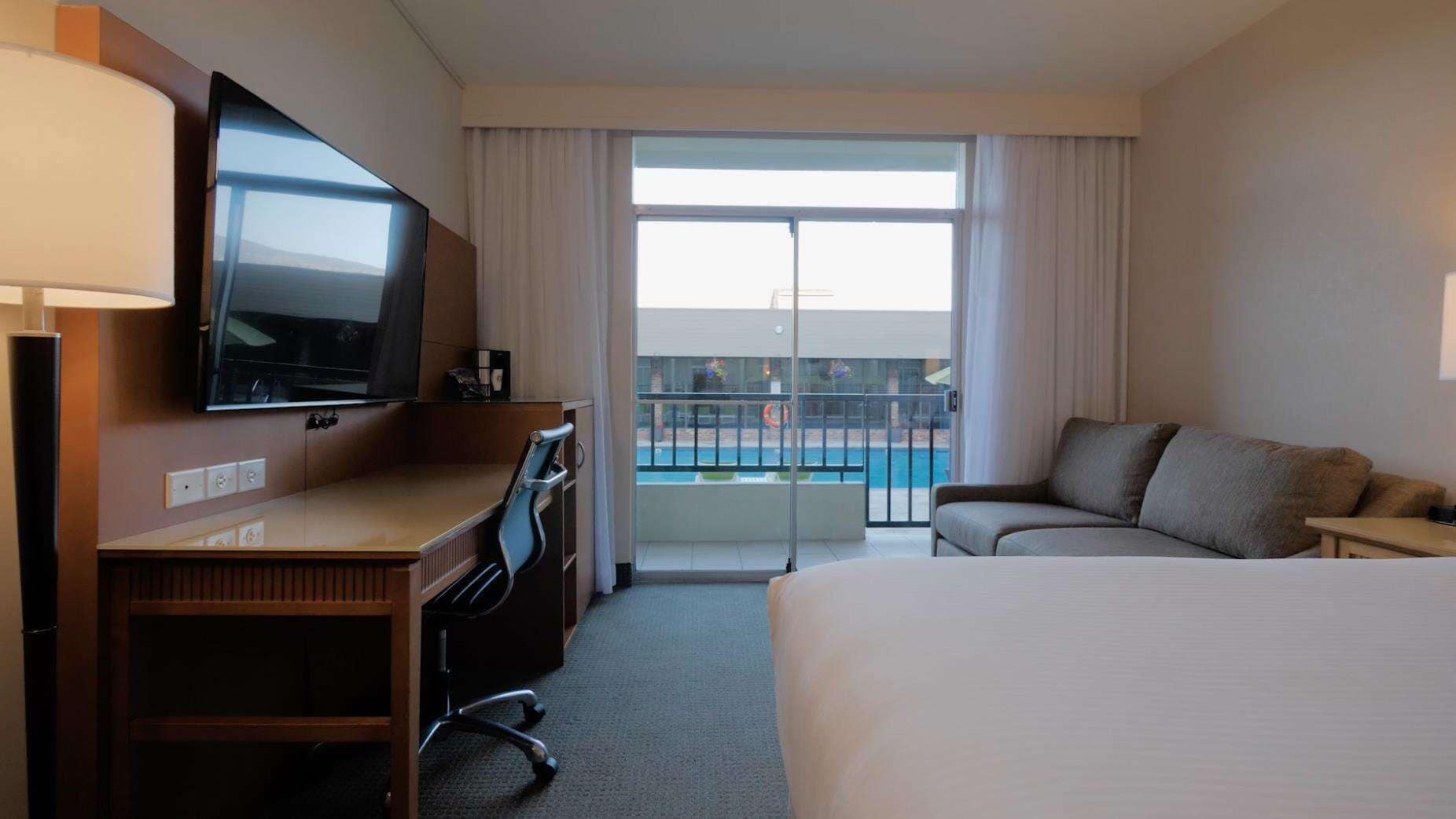 A hotel room with a bed, a desk with a TV, a sofa, and a balcony with a pool view.