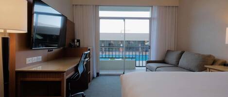 A hotel room with a bed, a desk with a TV, a sofa, and a balcony with a pool view.