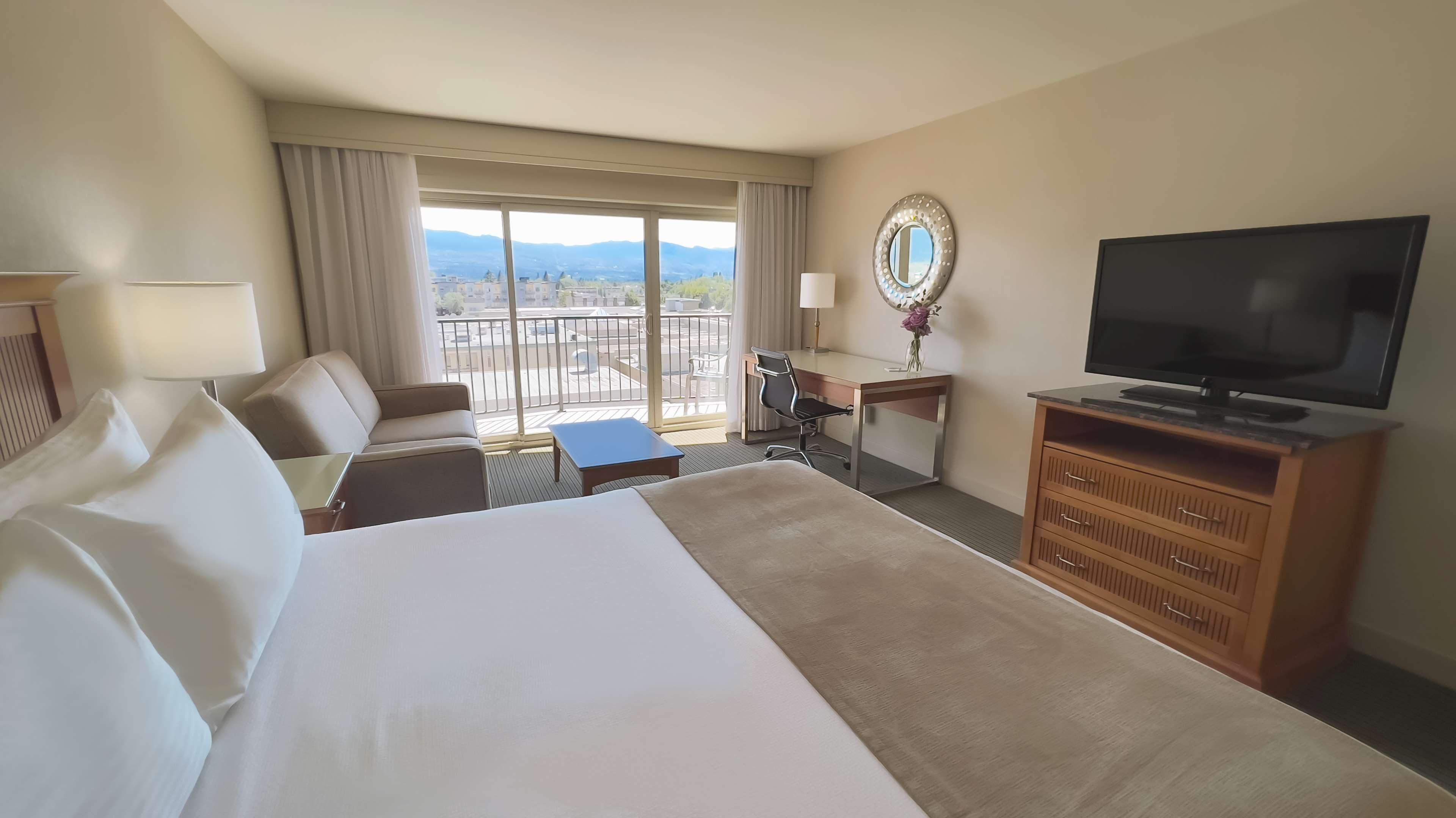 A hotel room with a large bed, a desk, a television, and a balcony with a view of the city.