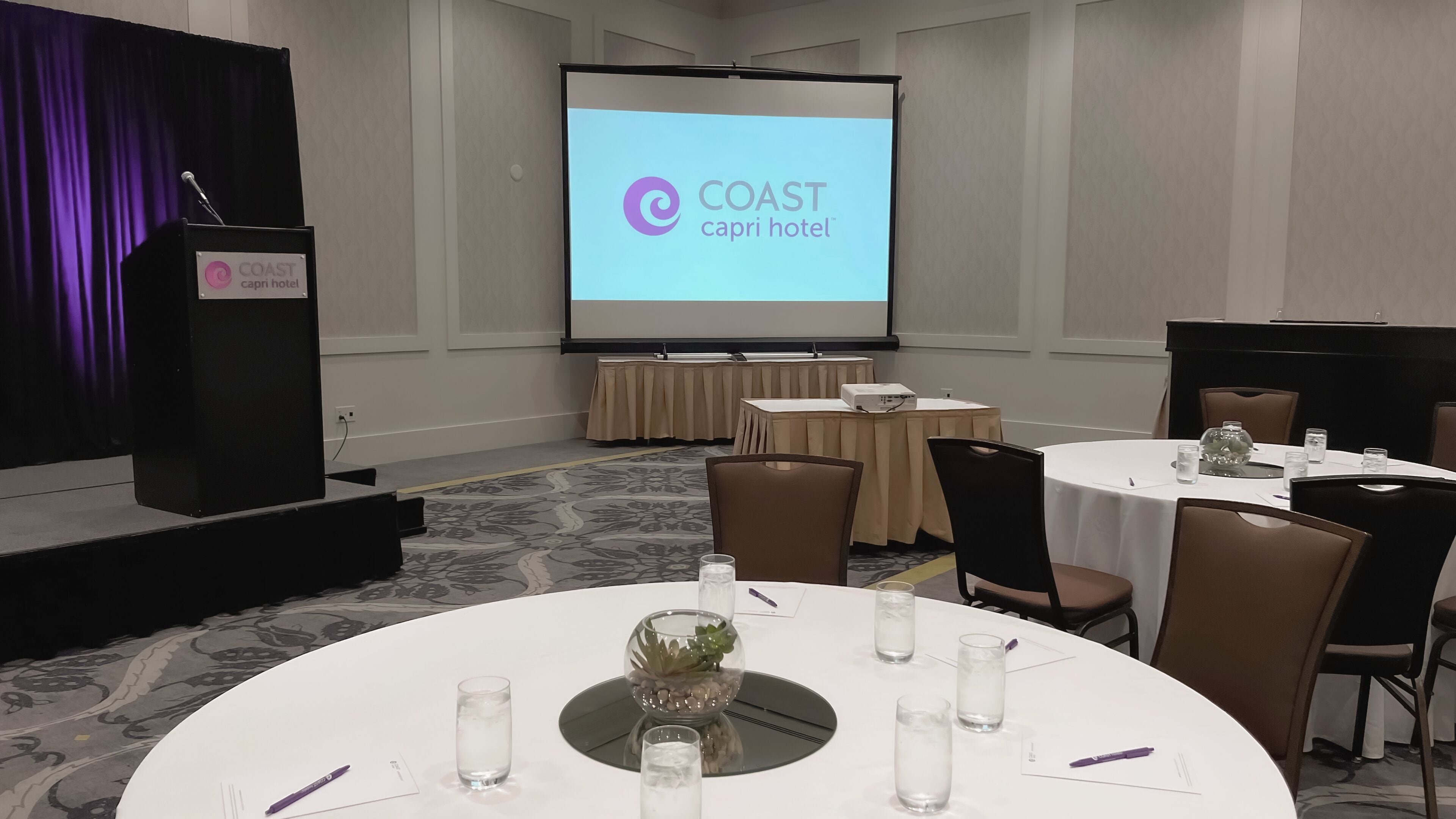 A conference room with a podium, a screen displaying the logo of COAST Capri Hotel, round tables with chairs, and a carpeted floor.