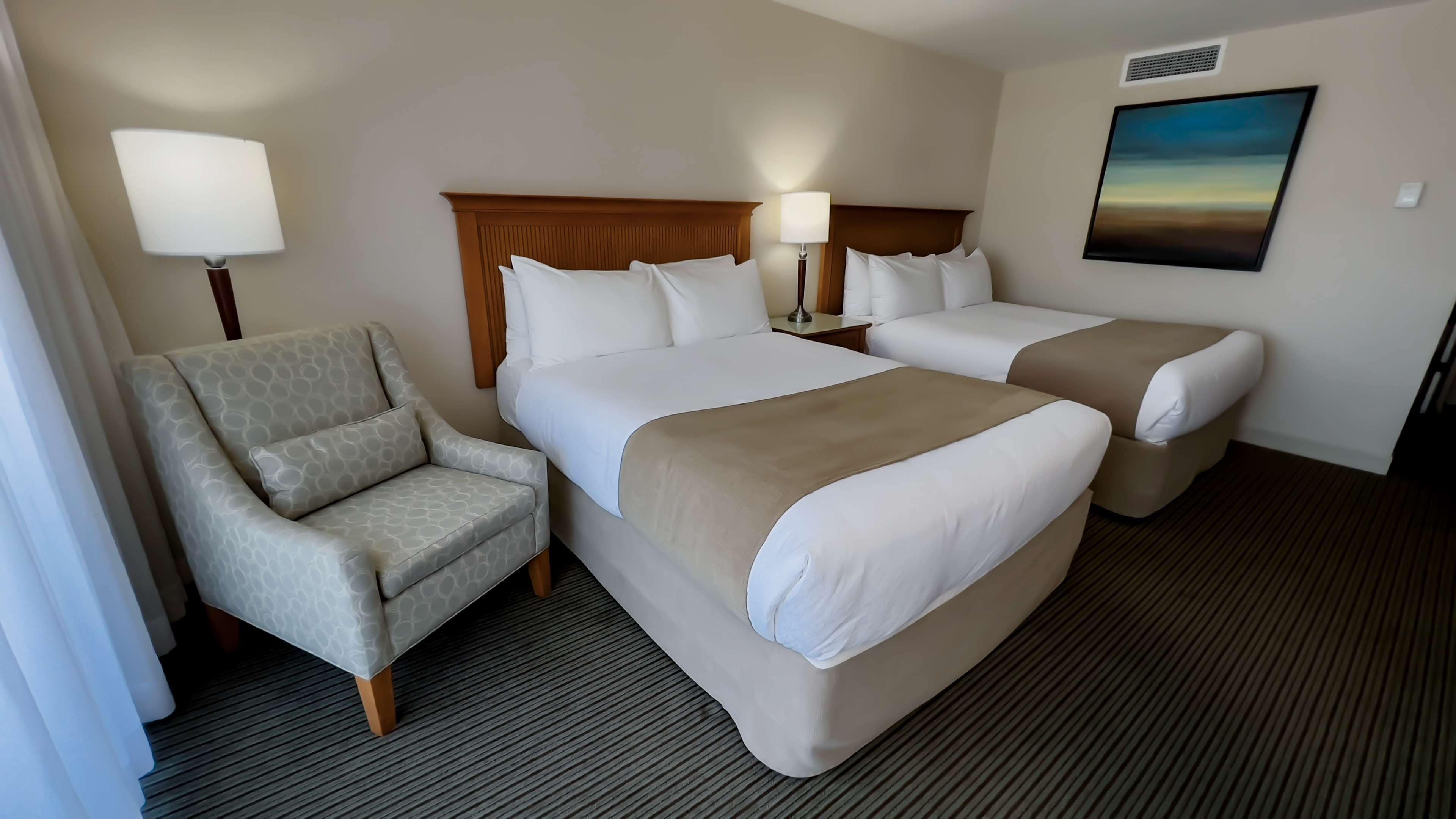 Habitación de hotel con dos camas, una silla, una lámpara y un cuadro en la pared.