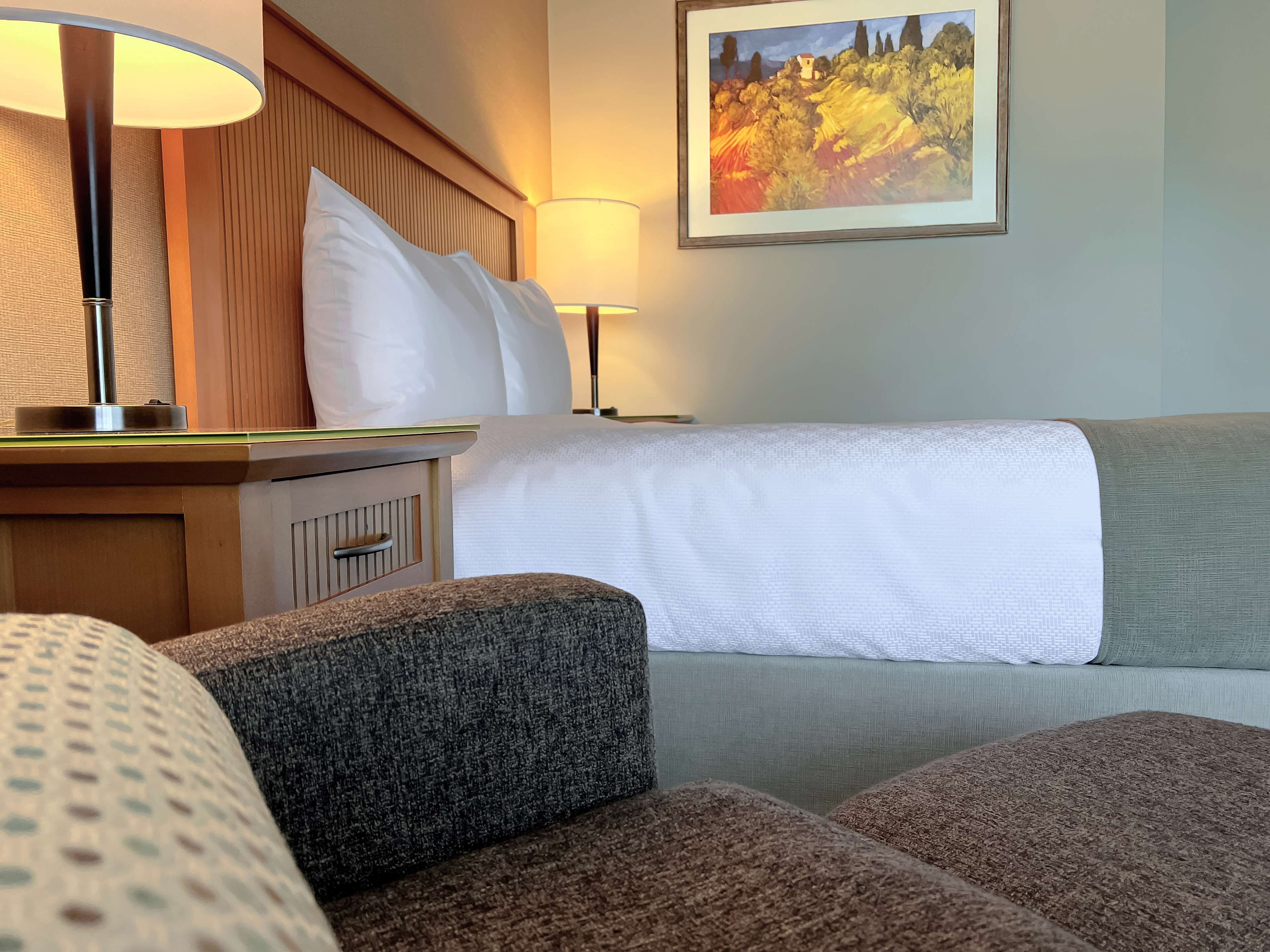 A hotel room with a bed, a nightstand with a lamp, a painting on the wall, and a sofa.