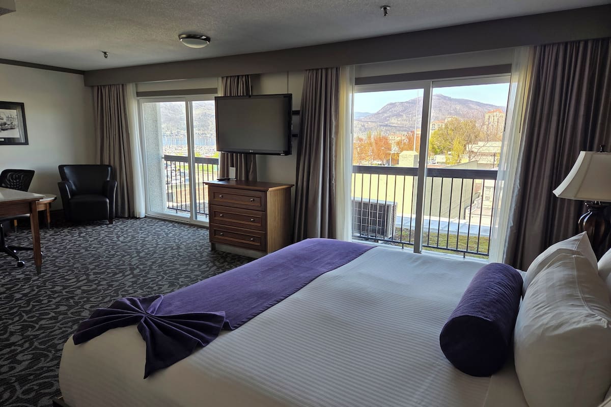 A hotel room with a large bed, a desk, a chair, a dresser, a TV, and a balcony with a view of the mountains.