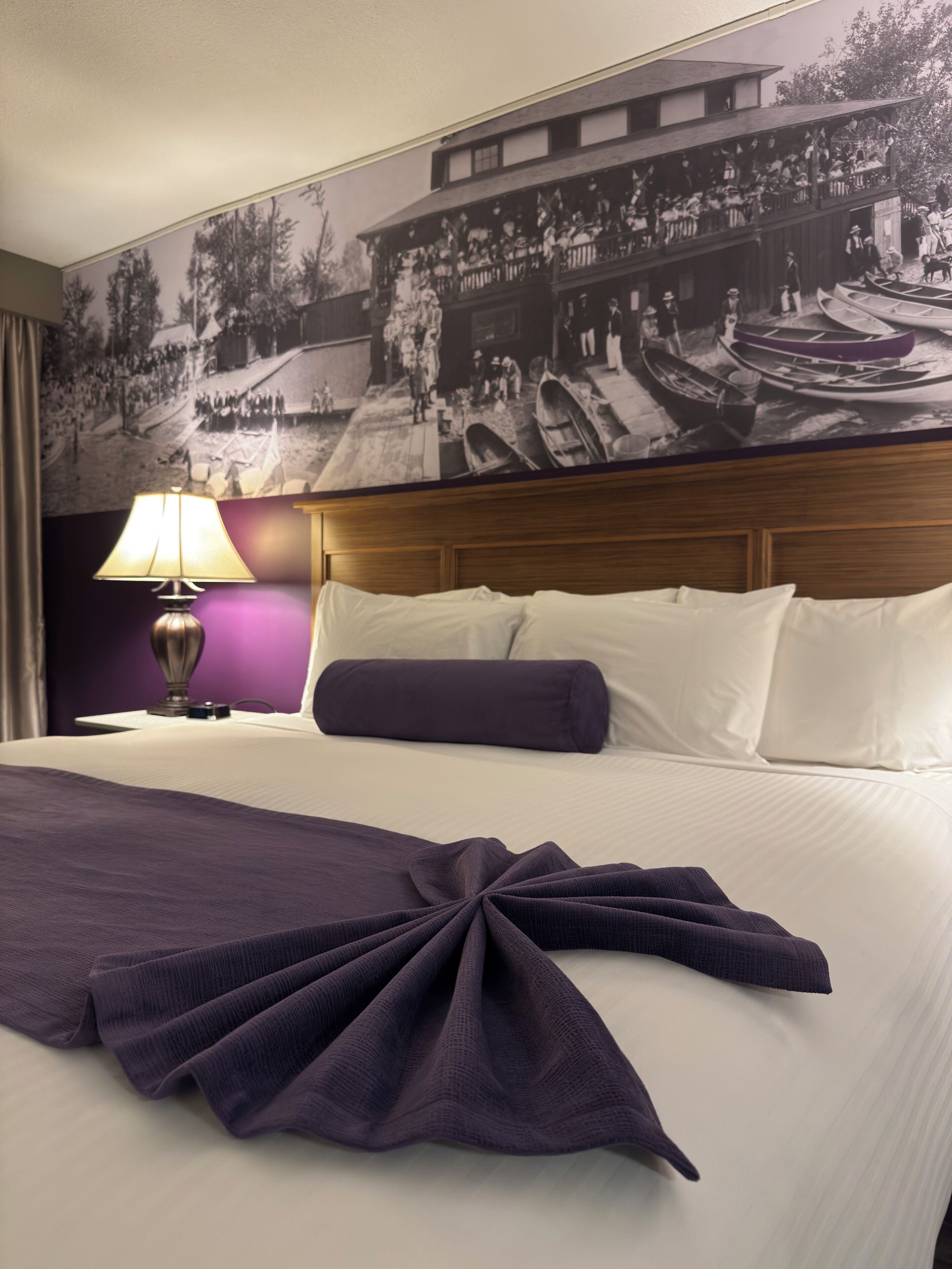 Habitación de hotel con una cama grande, cabecera, un cuadro colgado en la pared, una lámpara y un edredón doblado con cuidado.