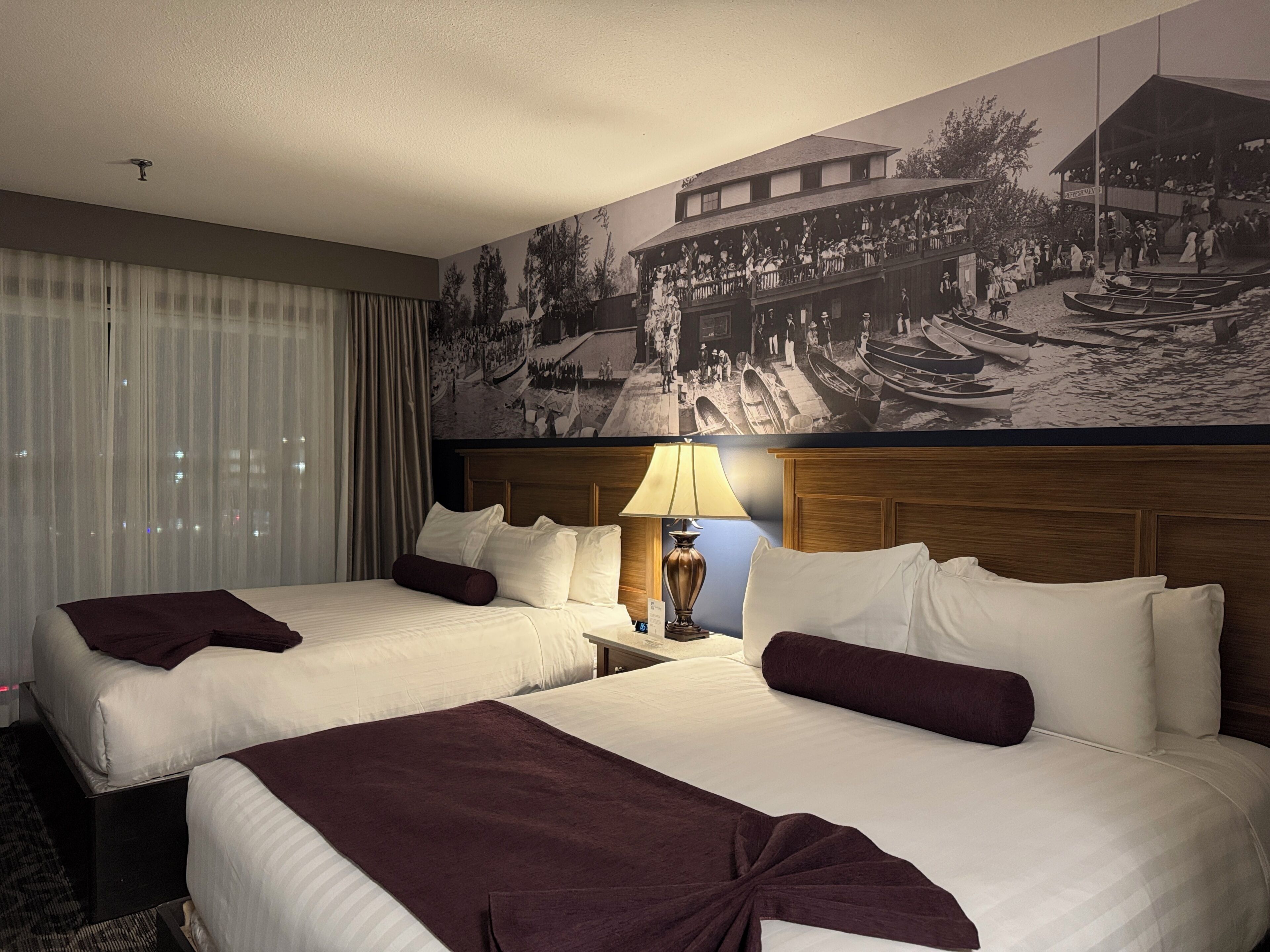 Ein Hotelzimmer mit zwei Betten, einem großen Wandbild, einem Fenster mit Vorhängen und einem Nachttisch mit einer Lampe.