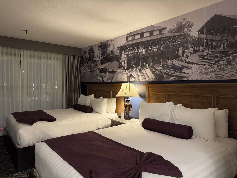 Ein Hotelzimmer mit zwei Betten, einem großen Wandbild, einem Fenster mit Vorhängen und einem Nachttisch mit einer Lampe.
