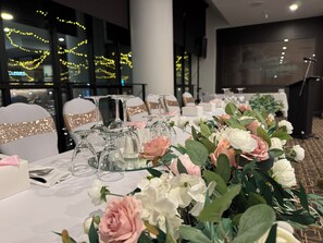 Una mesa de banquete dispuesta con cristalería y centros de flores, sillas con cintas decorativas y vista del interior con luces colgantes.