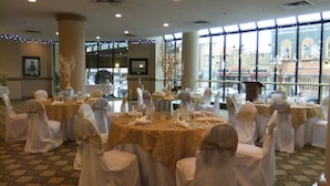 Un salón de banquetes con mesas redondas dispuestas para un evento formal, amplias ventanas con vistas a la ciudad y elementos decorativos como luces colgantes.
