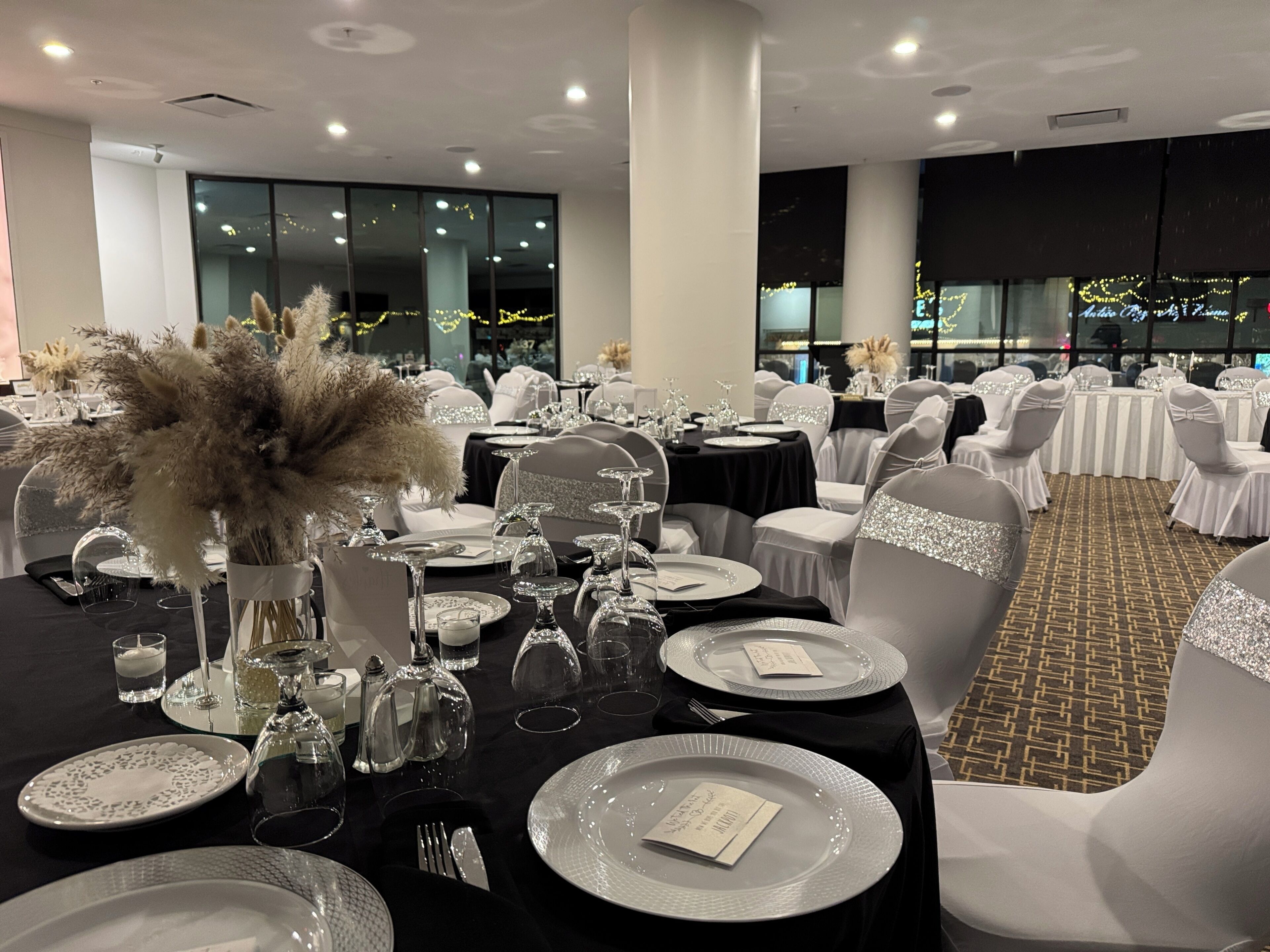 A banquet hall with round tables set for a formal event, featuring white chairs with glittery sashes, black tablecloths, and clear glassware.