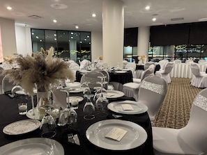 Un salón de banquetes con mesas redondas dispuestas para un evento formal, con sillas blancas adornadas con cintas brillantes, manteles negros y cristalería transparente.