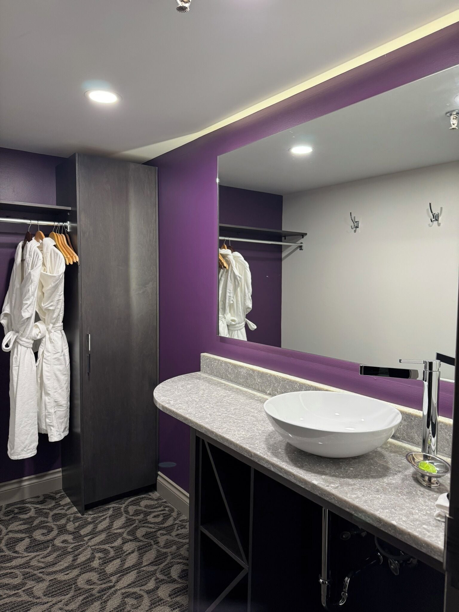 A modern bathroom with a large mirror, a white sink, and a robe rack with white robes.
