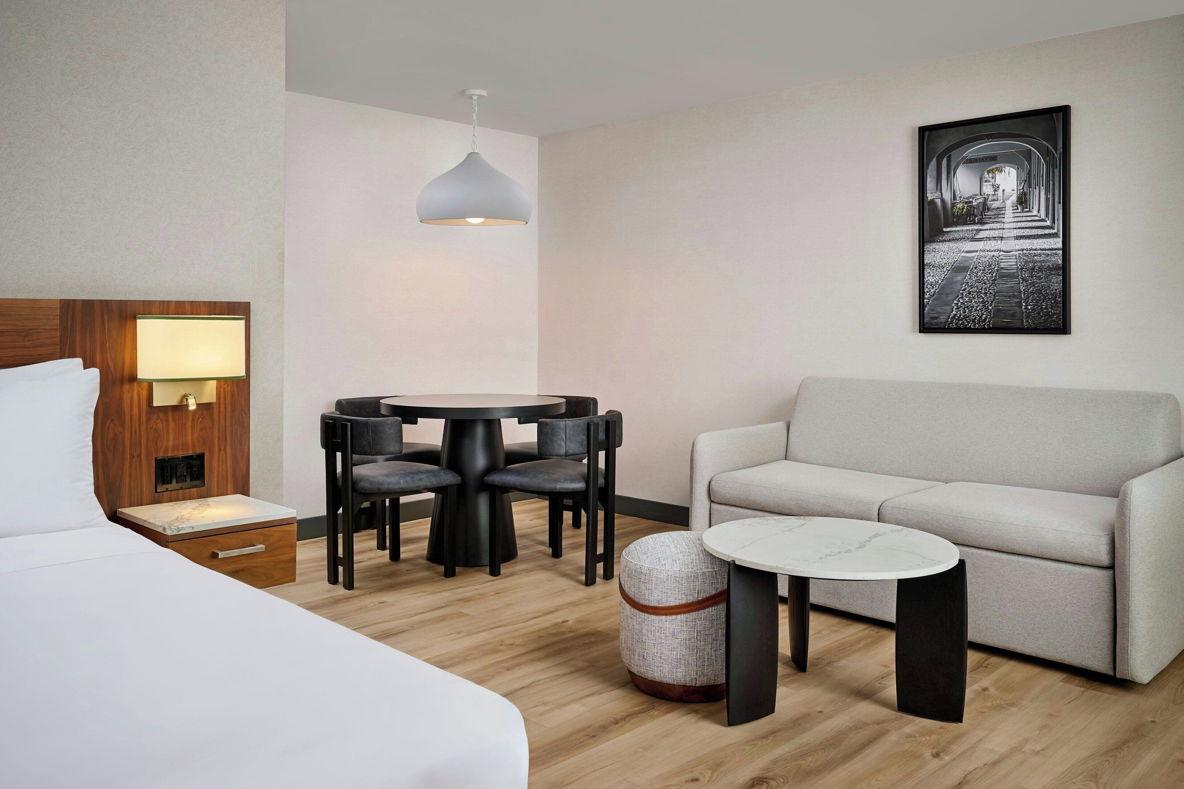 Habitación de hotel con cama, una mesa redonda con sillas, un sofá, una pequeña mesa redonda, una lámpara colgante y un cuadro en la pared.