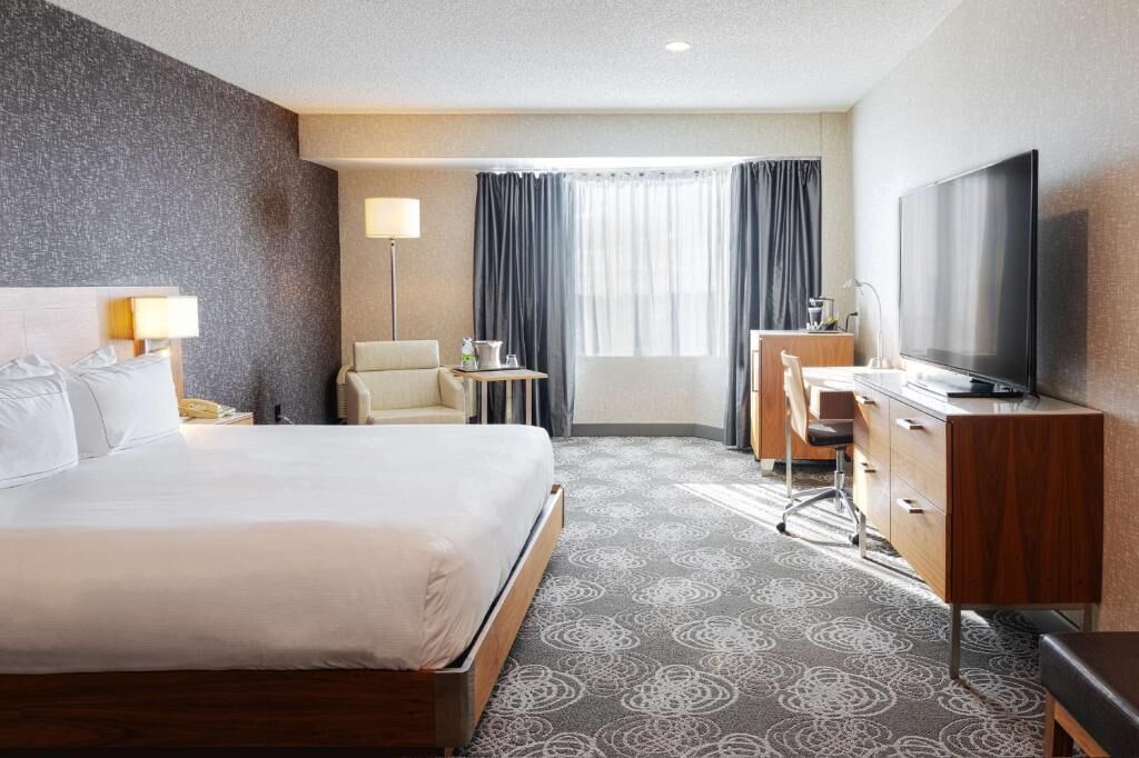 A hotel room with a bed, a desk, a television, a chair, a lamp, and a window with curtains.