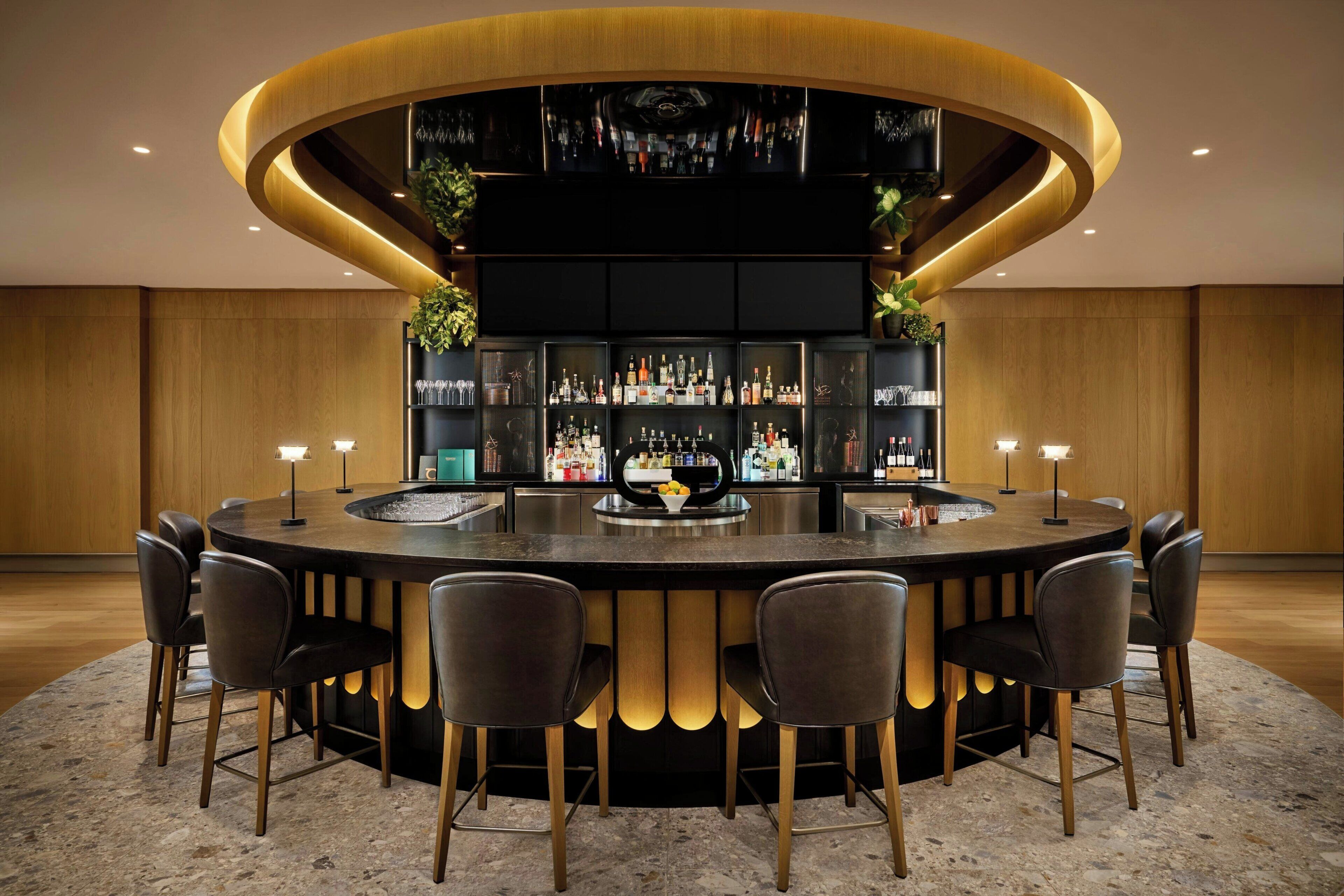 A circular bar area with a central bar, surrounded by six chairs, and shelves stocked with bottles and glasses.