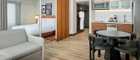 Una habitación de hotel con cama, sofá, mesa de comedor con sillas, y cocina equipada con microondas, cafetera y refrigerador.