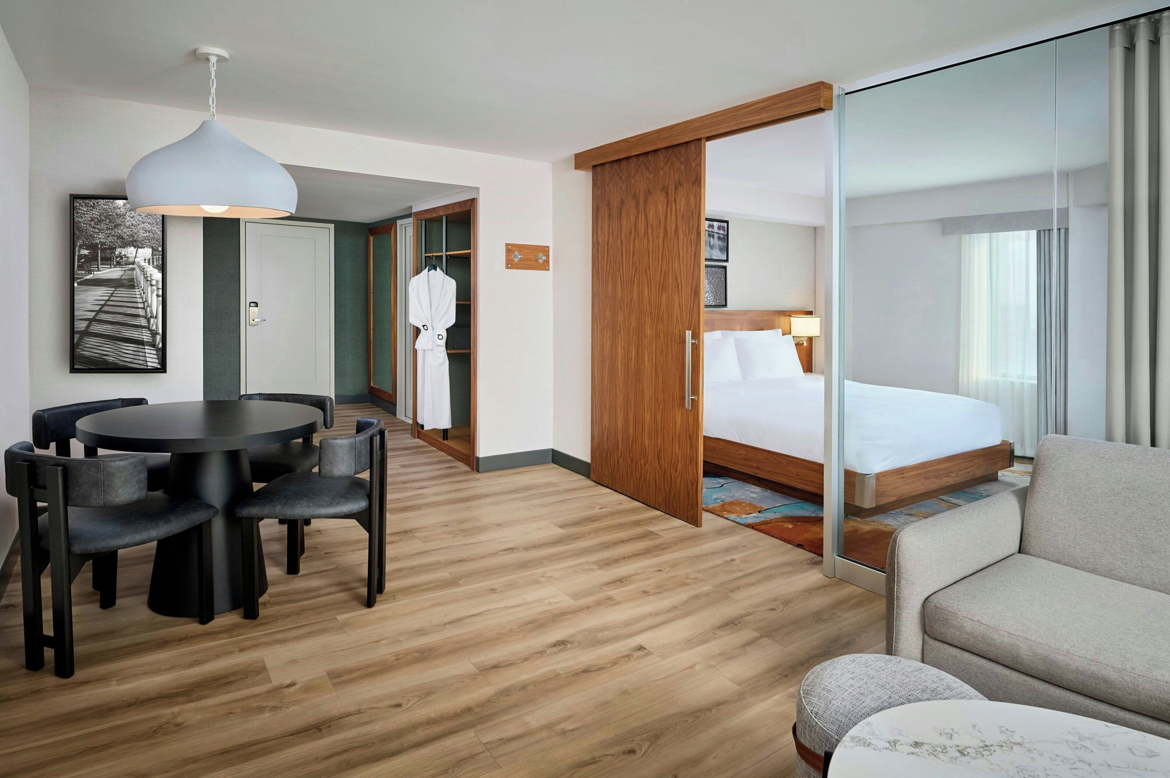 Habitación de hotel con cama, mesa redonda con sillas, sofá, mampara de vidrio y suelo de madera.