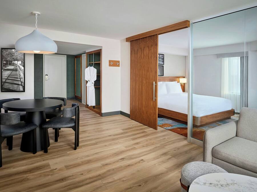 Habitación de hotel con cama, mesa redonda con sillas, sofá, mampara de vidrio y suelo de madera.