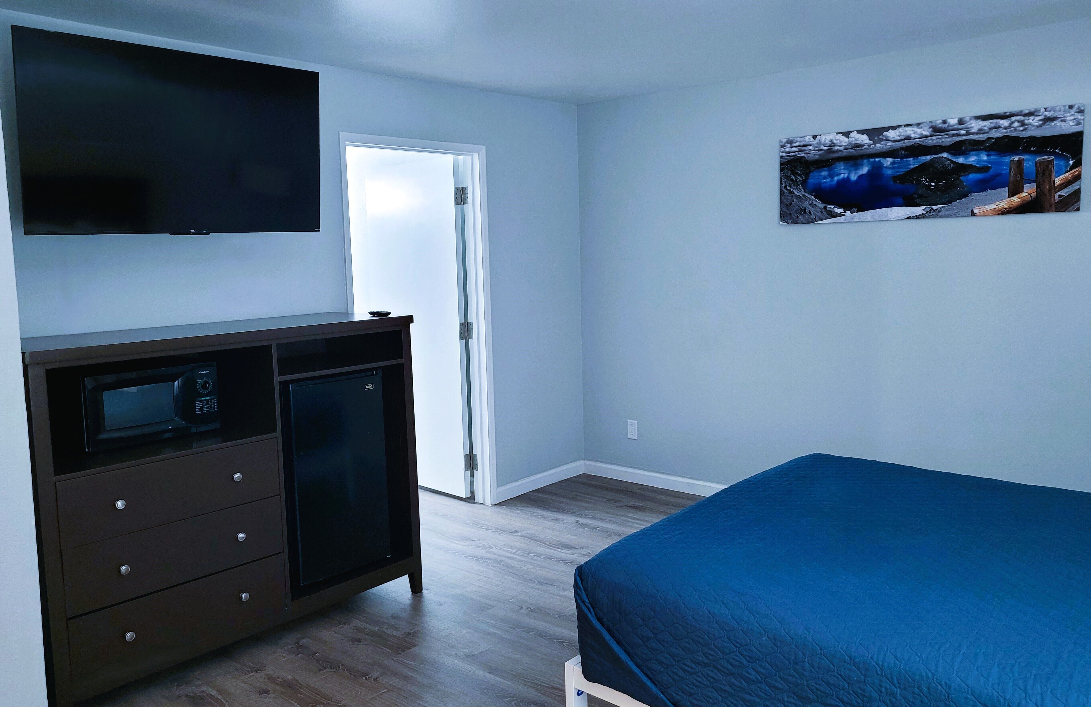 Un dormitorio con una cama, un televisor montado en la pared, un mueble bajo con cajones y una puerta que da a otra habitación.