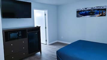 A bedroom with a bed, a television mounted on the wall, a small cabinet with drawers, and a door leading to another room.