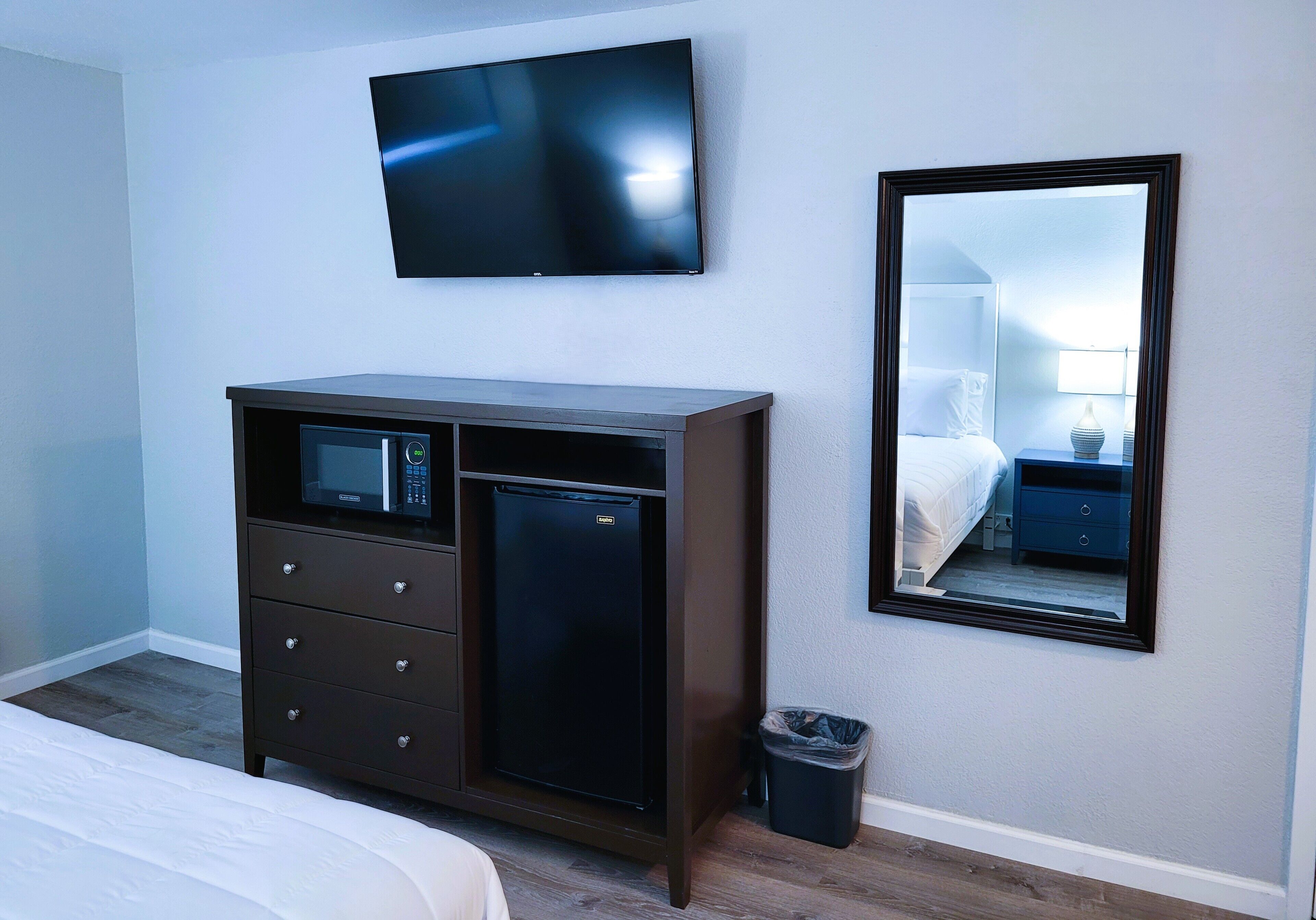 A hotel room with a bed, a dresser with a microwave, a TV, a mirror, and a trash can.
