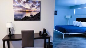 Ein Zimmer mit einem Bett, einem Schreibtisch mit Stuhl, einer Lampe, einer Kaffeemaschine und einem Bild eines Sonnenuntergangs über dem Wasser.