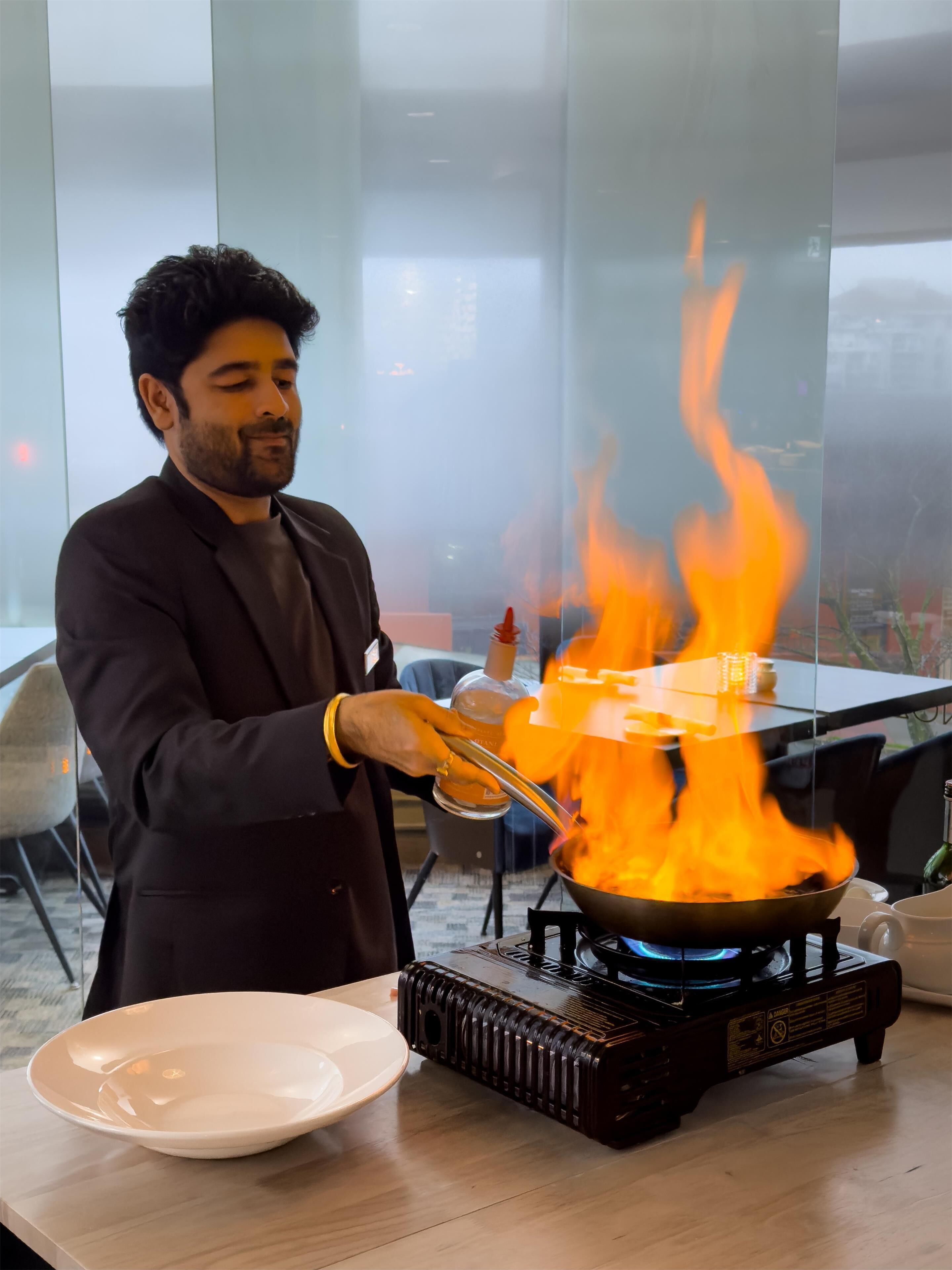 A person cooking with a pan on a portable stove, surrounded by a dining area with chairs and a table.