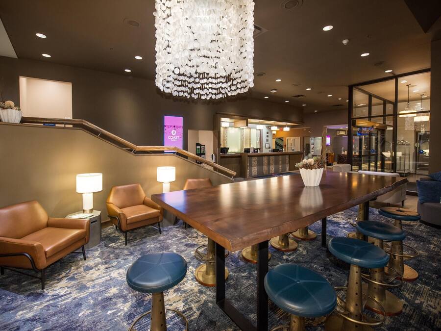 A hotel lobby with a wooden reception desk, blue bar stools, leather armchairs, and a chandelier.