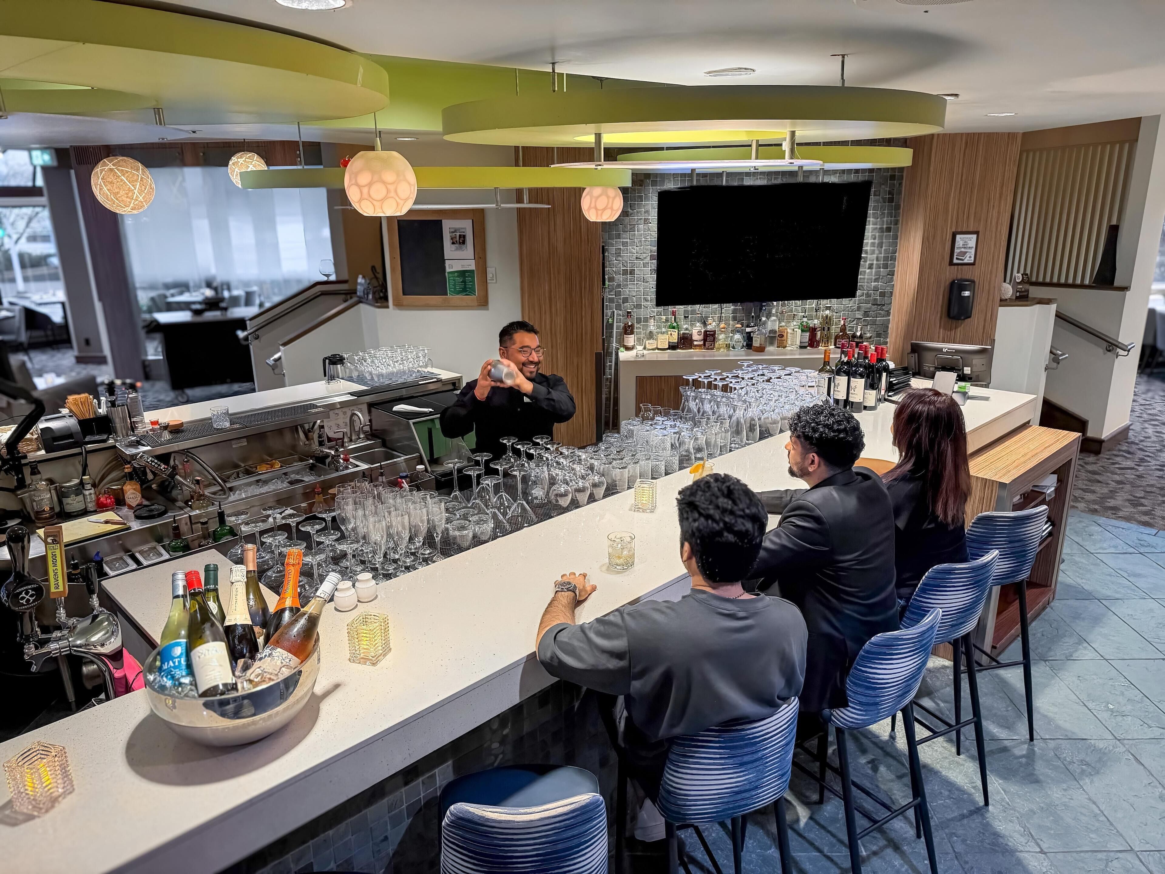 En modern bardisk med en bartender som blandar drinkar, en disk med olika flaskor och glas, och en sittgrupp med barstolar.