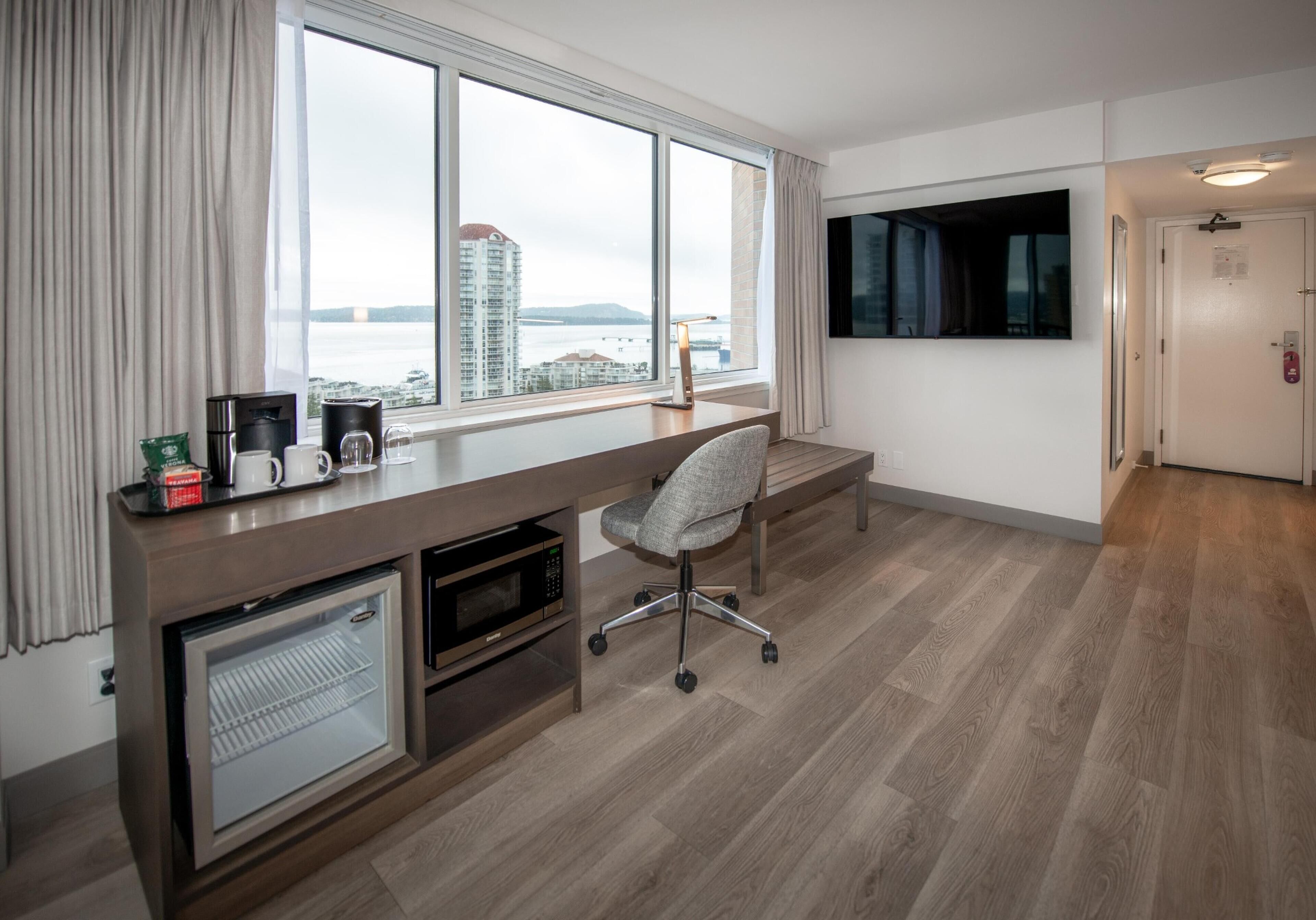 A modern hotel room with a large window, a desk with a chair, a mini-fridge, a microwave, and a flat-screen TV.
