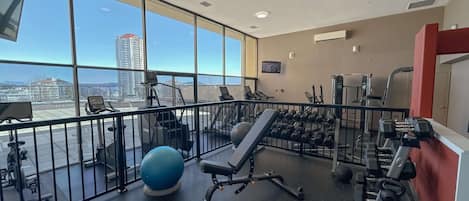 Un gimnasio con equipo de ejercicio, una pelota azul para hacer ejercicio y vistas de un paisaje urbano a través de amplias ventanas.