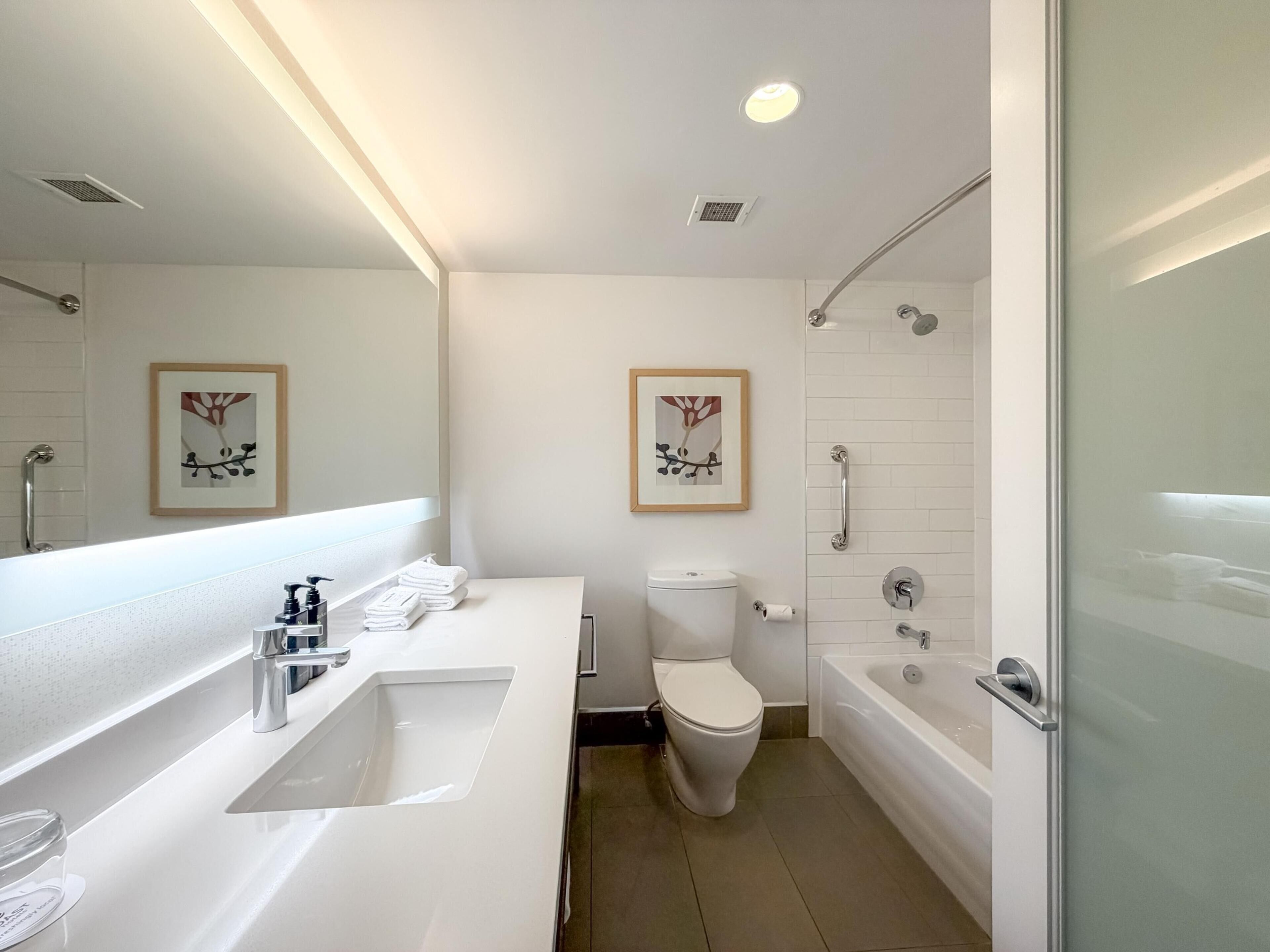 Ein modernes Badezimmer mit einem weißen Waschbecken, einer Toilette und einer Badewanne. An den Wänden hängen zwei gerahmte Bilder, und das Zimmer ist in einem klaren, minimalistischen Stil gehalten.