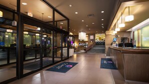 Un vestíbulo de hotel con mostrador de recepción, puertas de cristal y una escalera que sube al piso superior.