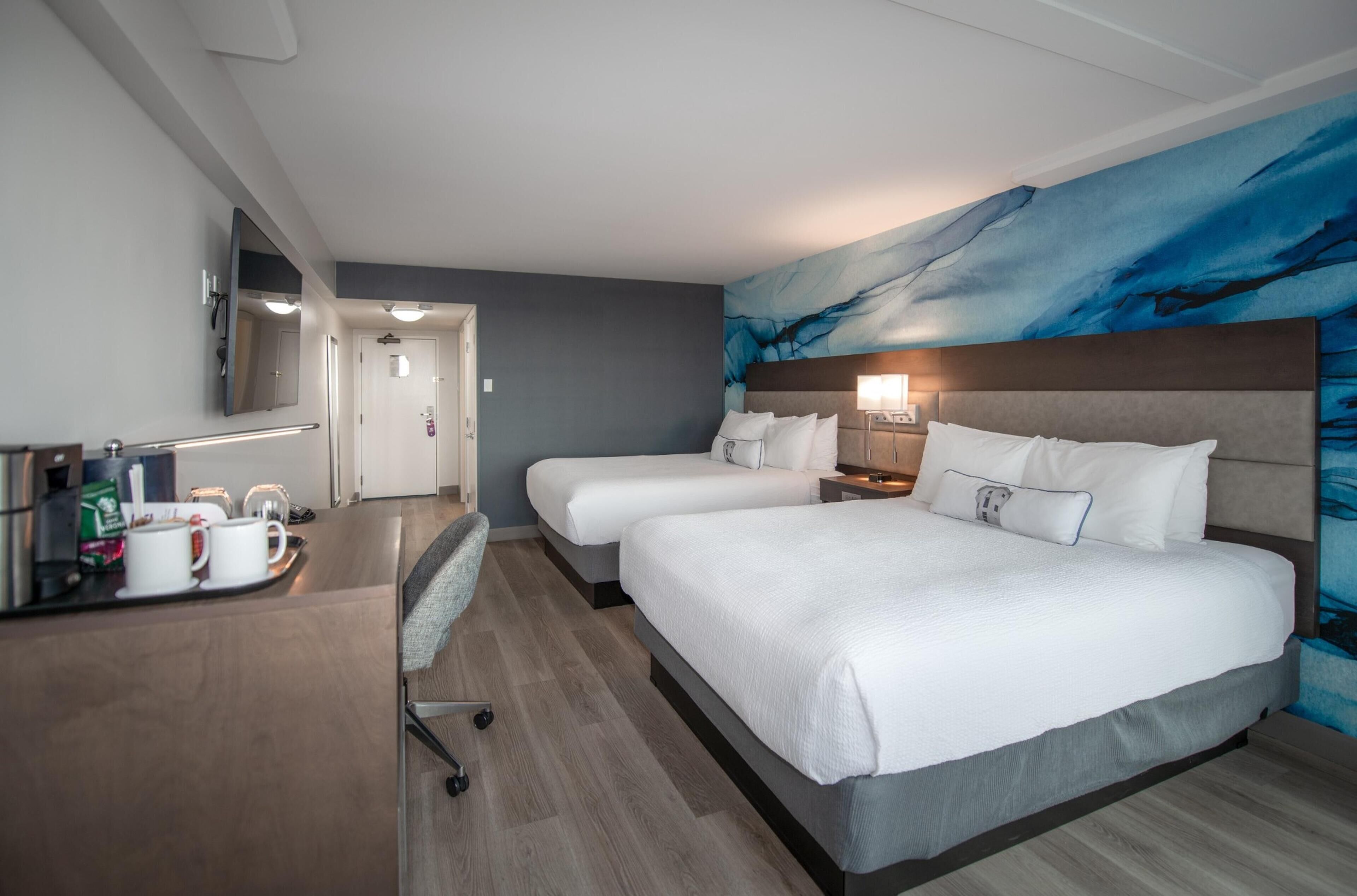 A hotel room with two beds, a desk with a coffee maker, and a large abstract mural on the wall.