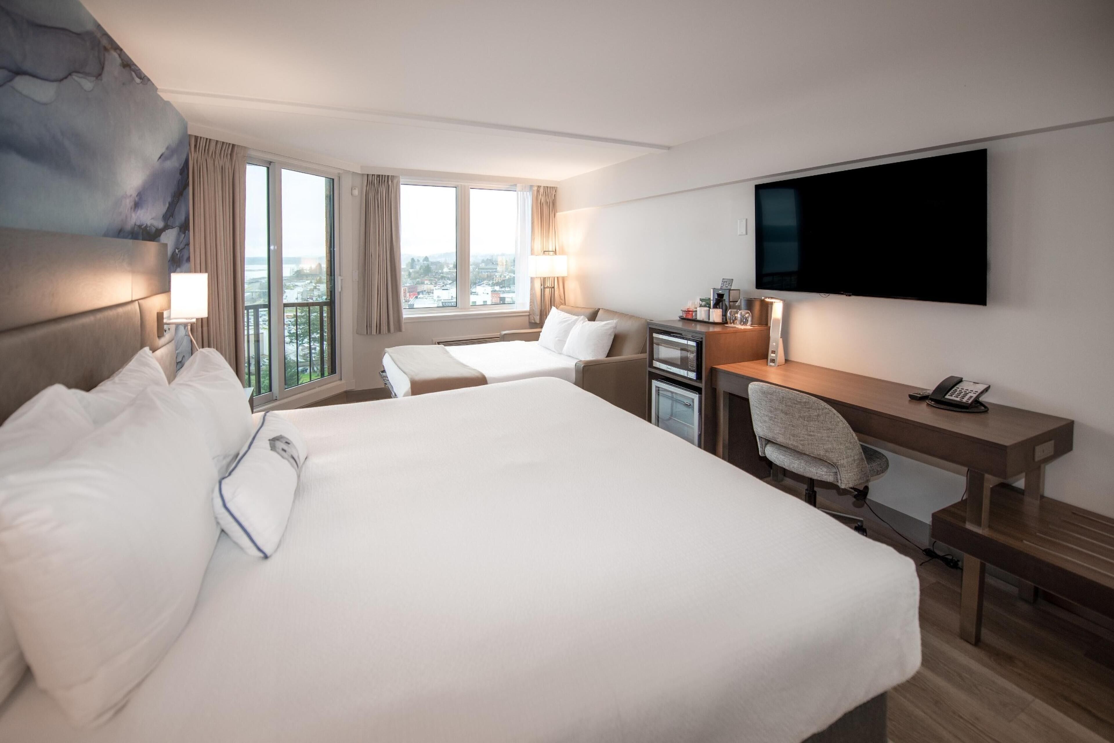 A hotel room with a large bed, a desk with a chair, a flat-screen TV, and a view of the city through large windows.