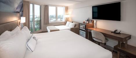 A hotel room with a large bed, a desk with a chair, a flat-screen TV, and a view of the city through large windows.