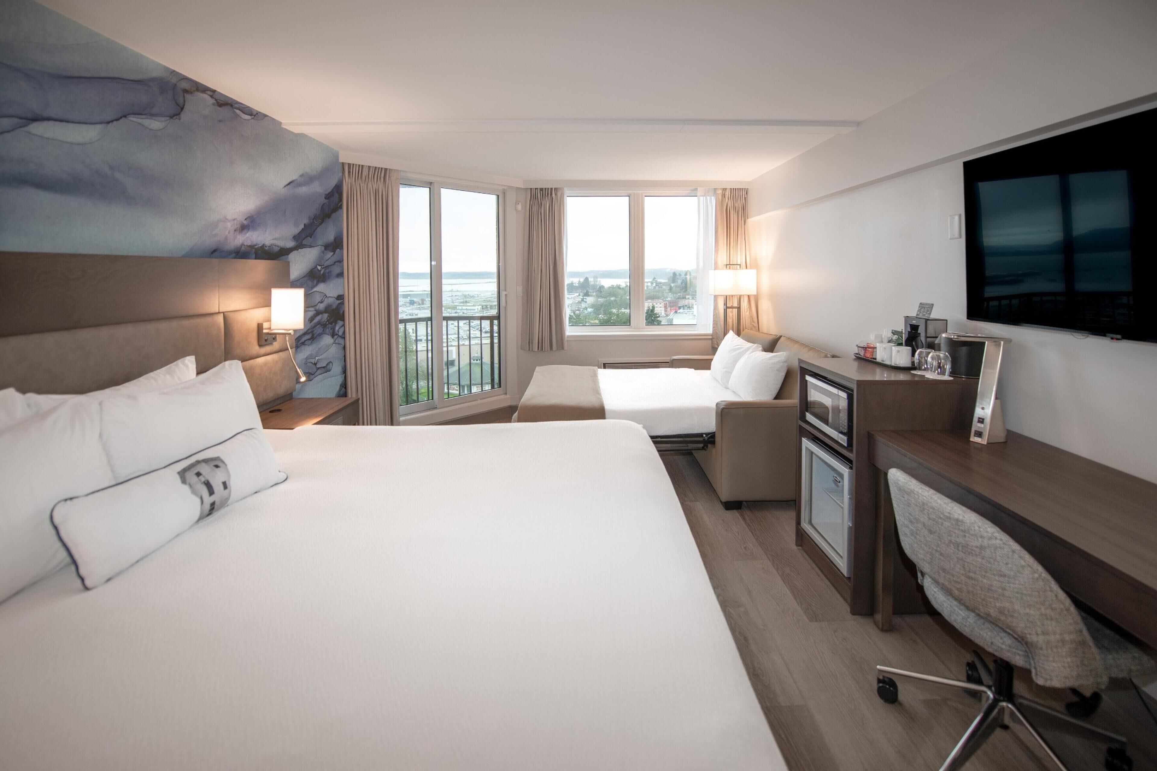 A hotel room with a large bed, a desk with a chair, a TV, and a view of the city.