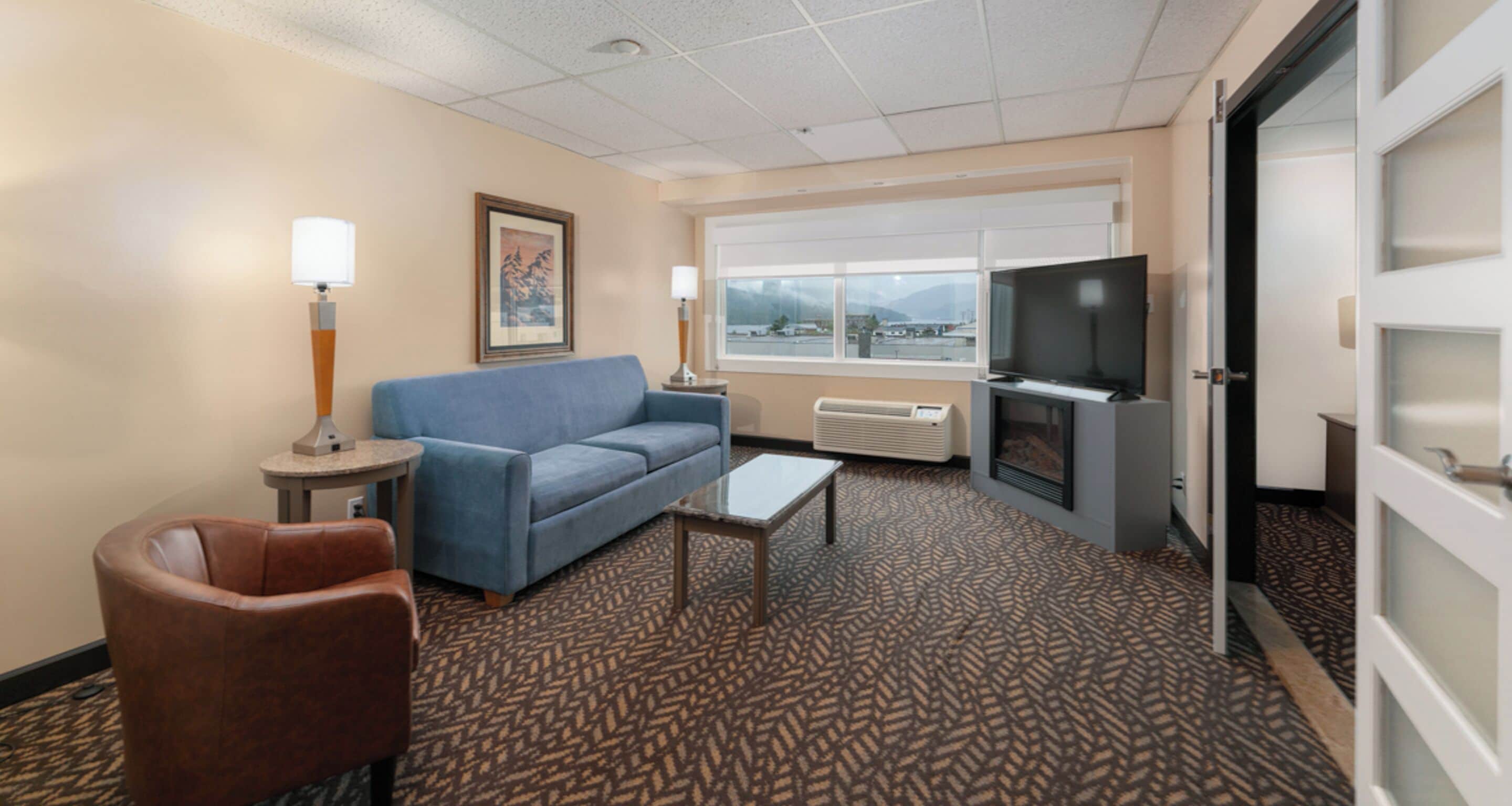 Habitación de hotel con sofá azul, sillón marrón, una mesita, televisor y ventana con vistas a montañas.