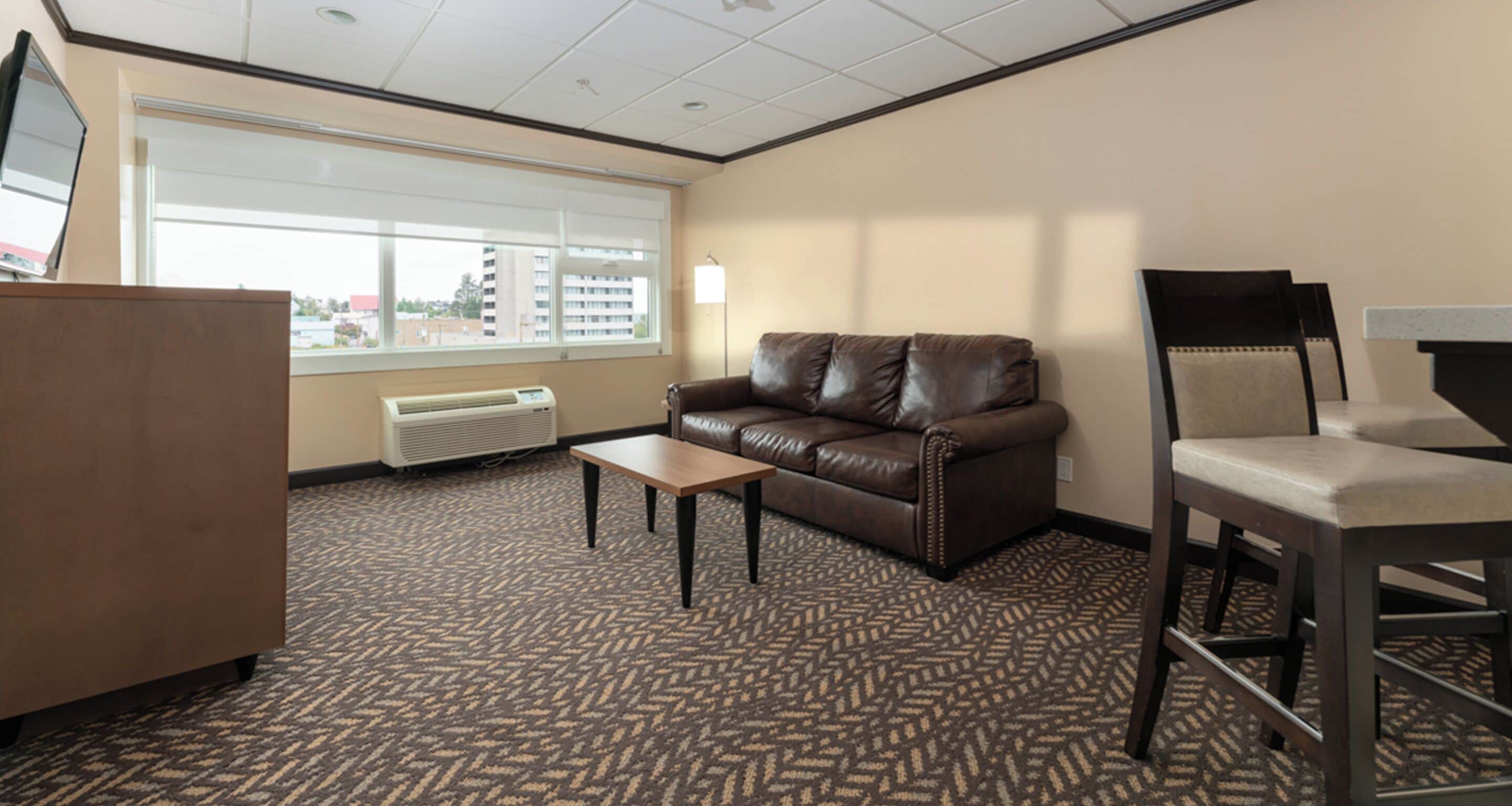 A hotel room with a brown leather sofa, a wooden desk, and a chair. There is a window with blinds, a radiator, and a patterned carpet.